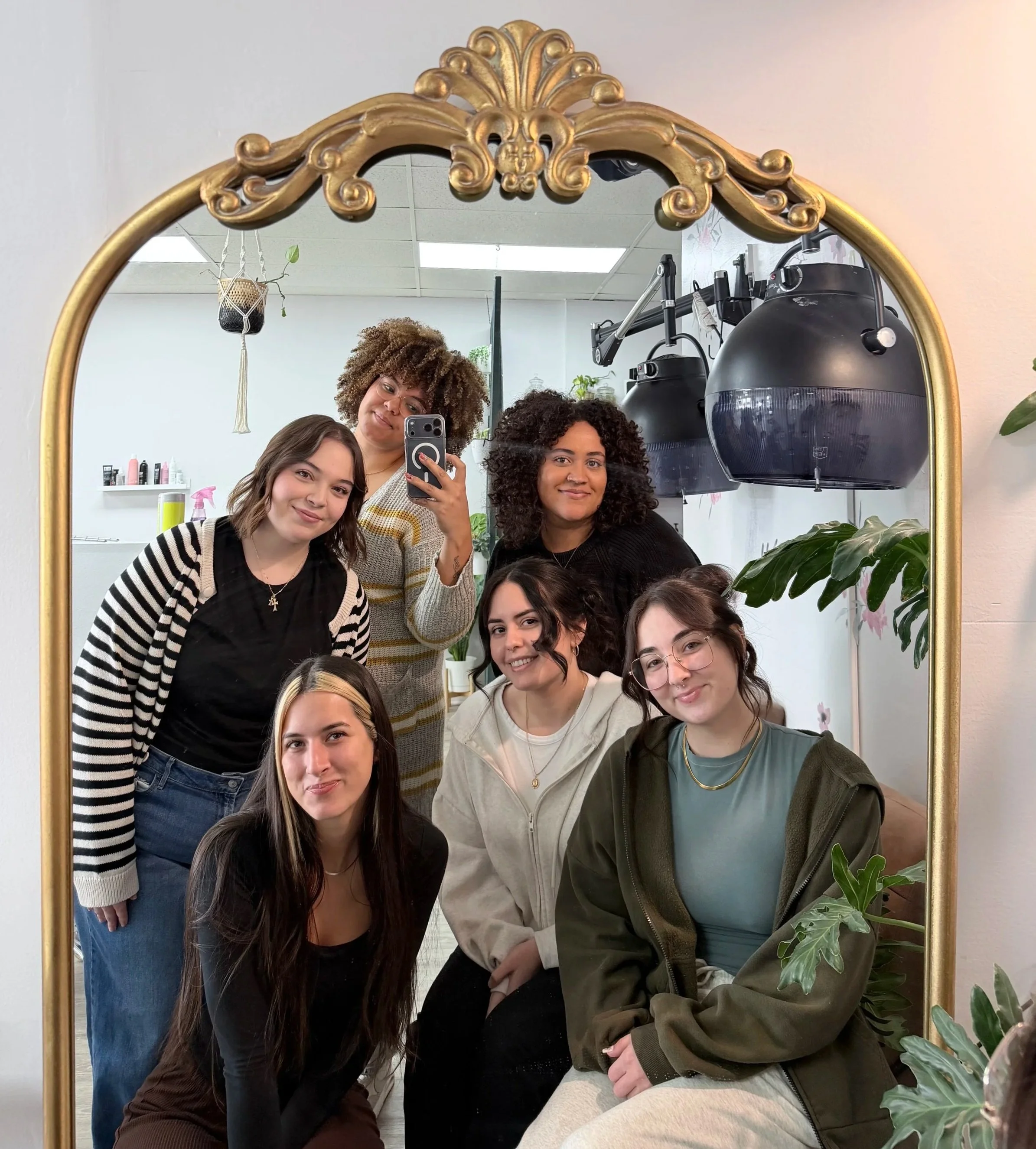Six women with diverse hair styles and colors posing for a mirror selfie in a hair salon, with salon equipment in the background and plants nearby.