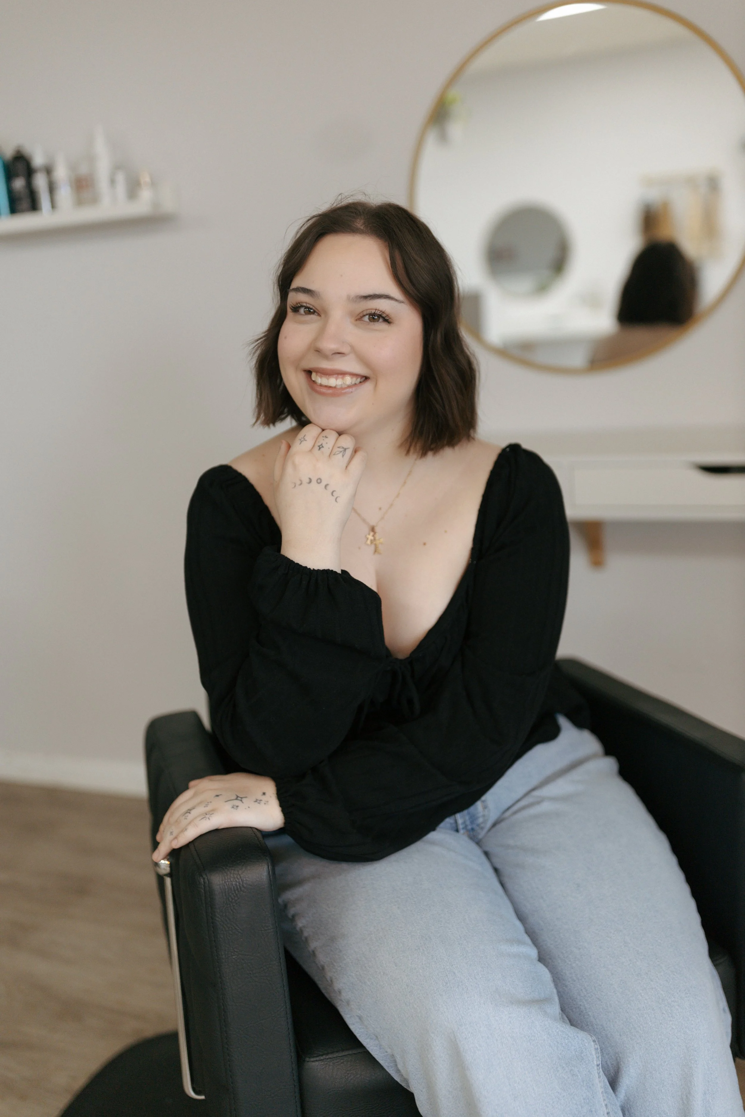 A young woman with long brown hair and light skin, smiling and sitting on a beige sofa with a large green leafy plant behind her, wearing a black long-sleeve top, a denim vest, and layered gold necklaces.