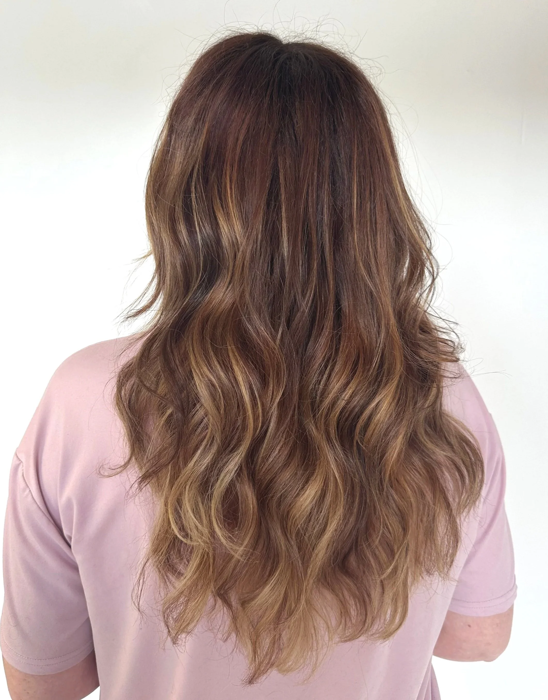 Woman with long, wavy, honey-blonde hair, seen from behind, wearing a light pink shirt.