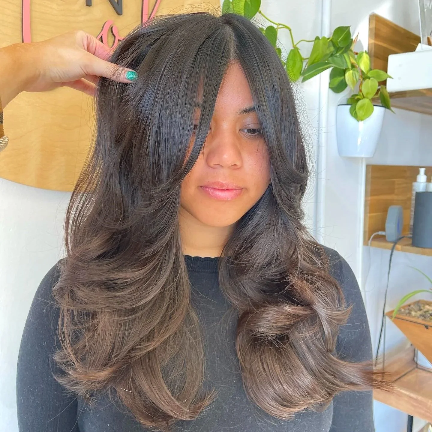 A woman with long, wavy hair having highlights, sitting in a salon, with a hand touching her hair.
