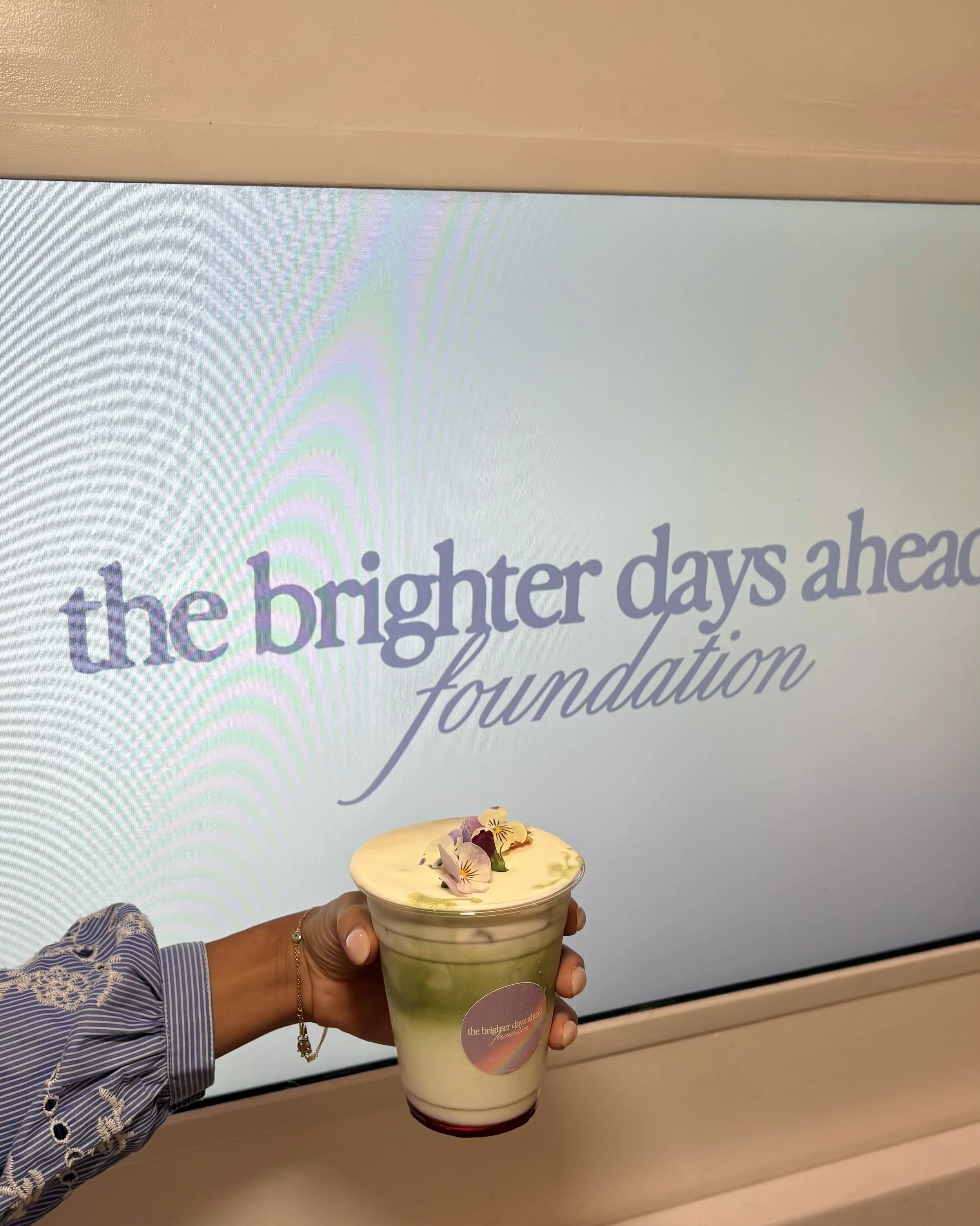 Iced matcha latte topped with edible flowers, held in front of a branded screen for The Brighter Days Ahead Foundation
