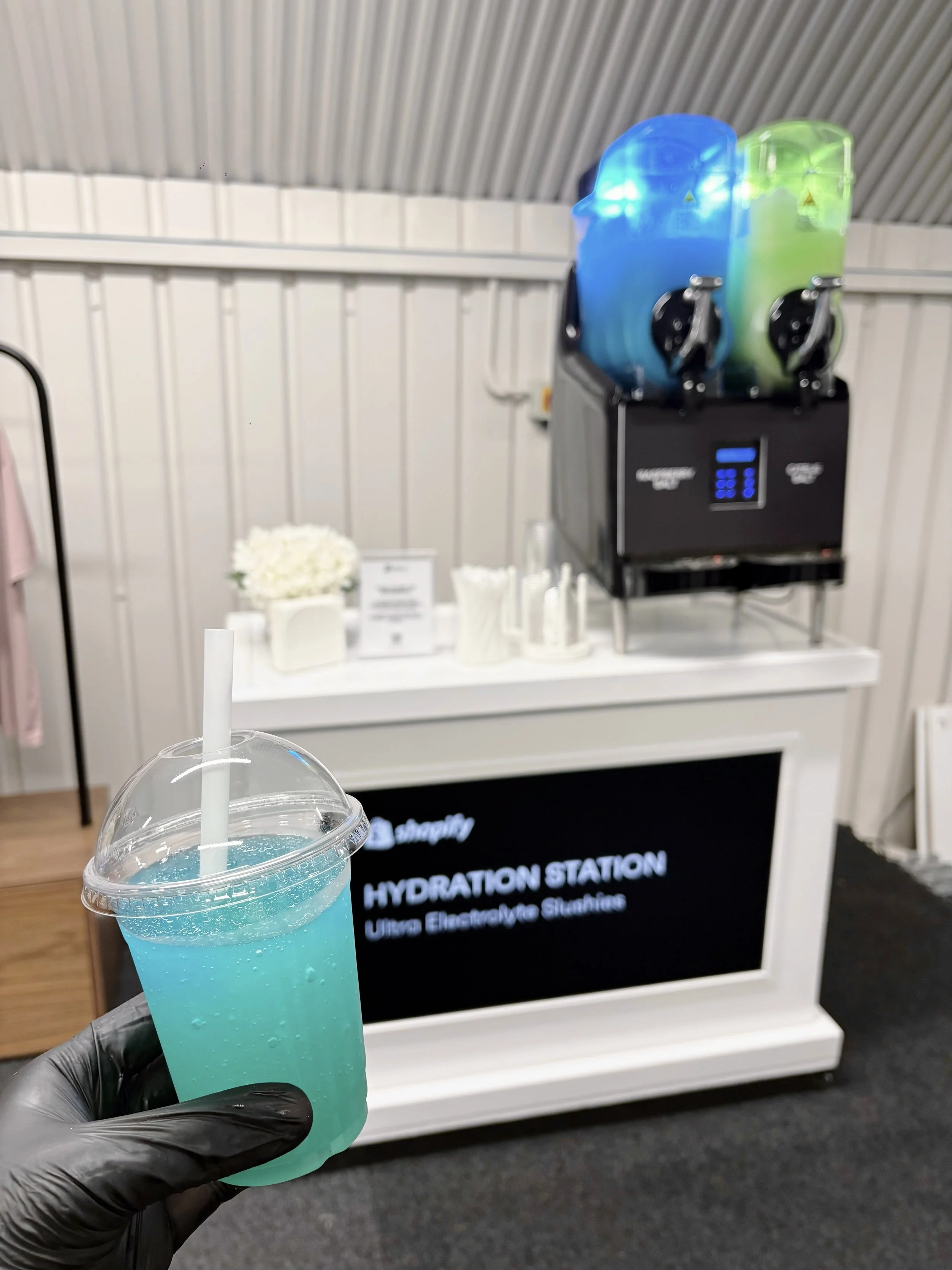 Blue electrolyte slush drink in a clear cup held in front of a hydration station bar, with twin slush machines filled with blue and green drinks in the background.