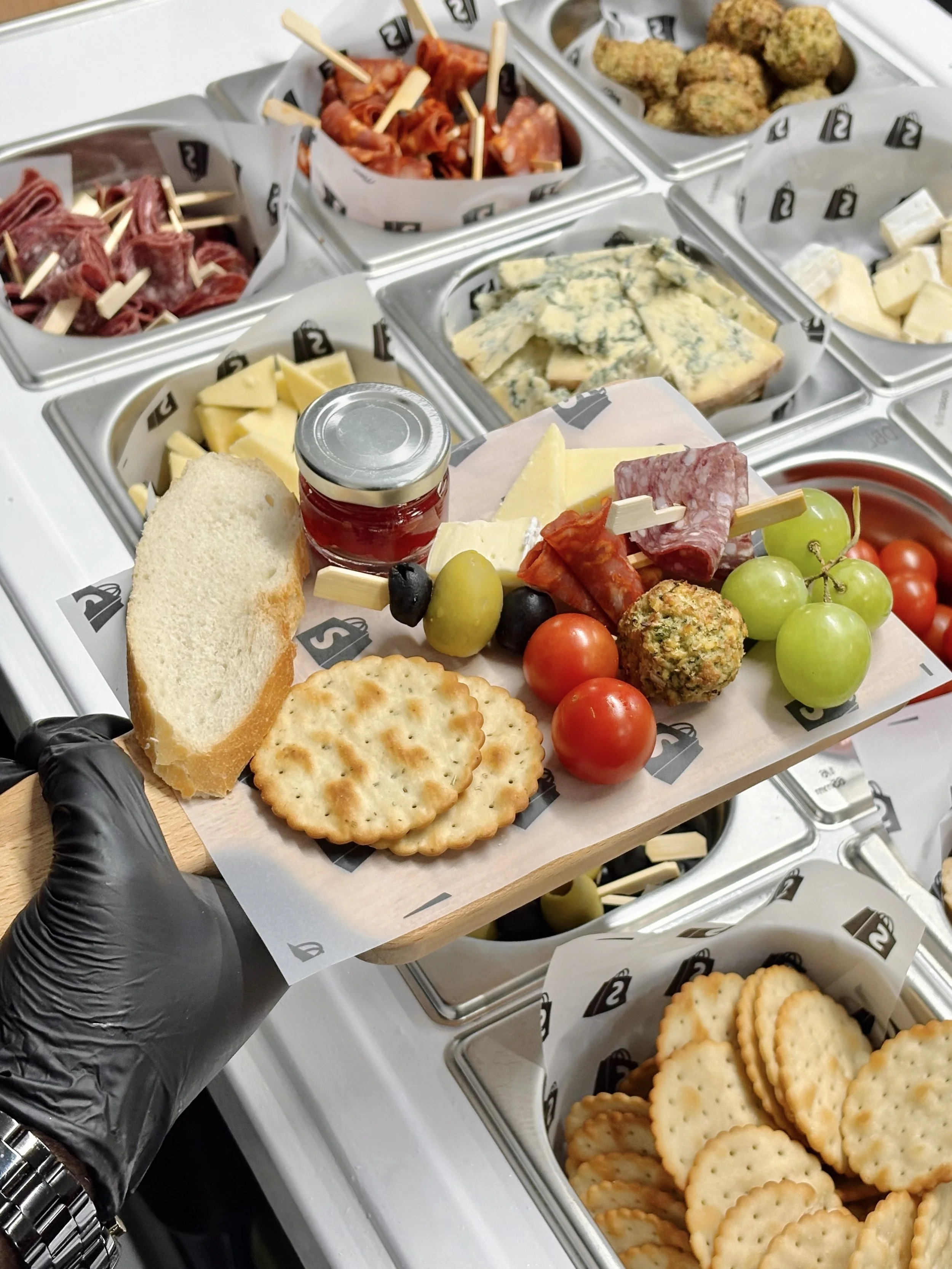 Gloved hand holding a charcuterie board above a chilled display of cheeses, meats, crackers, and olives, showcasing a premium grazing setup at an event.