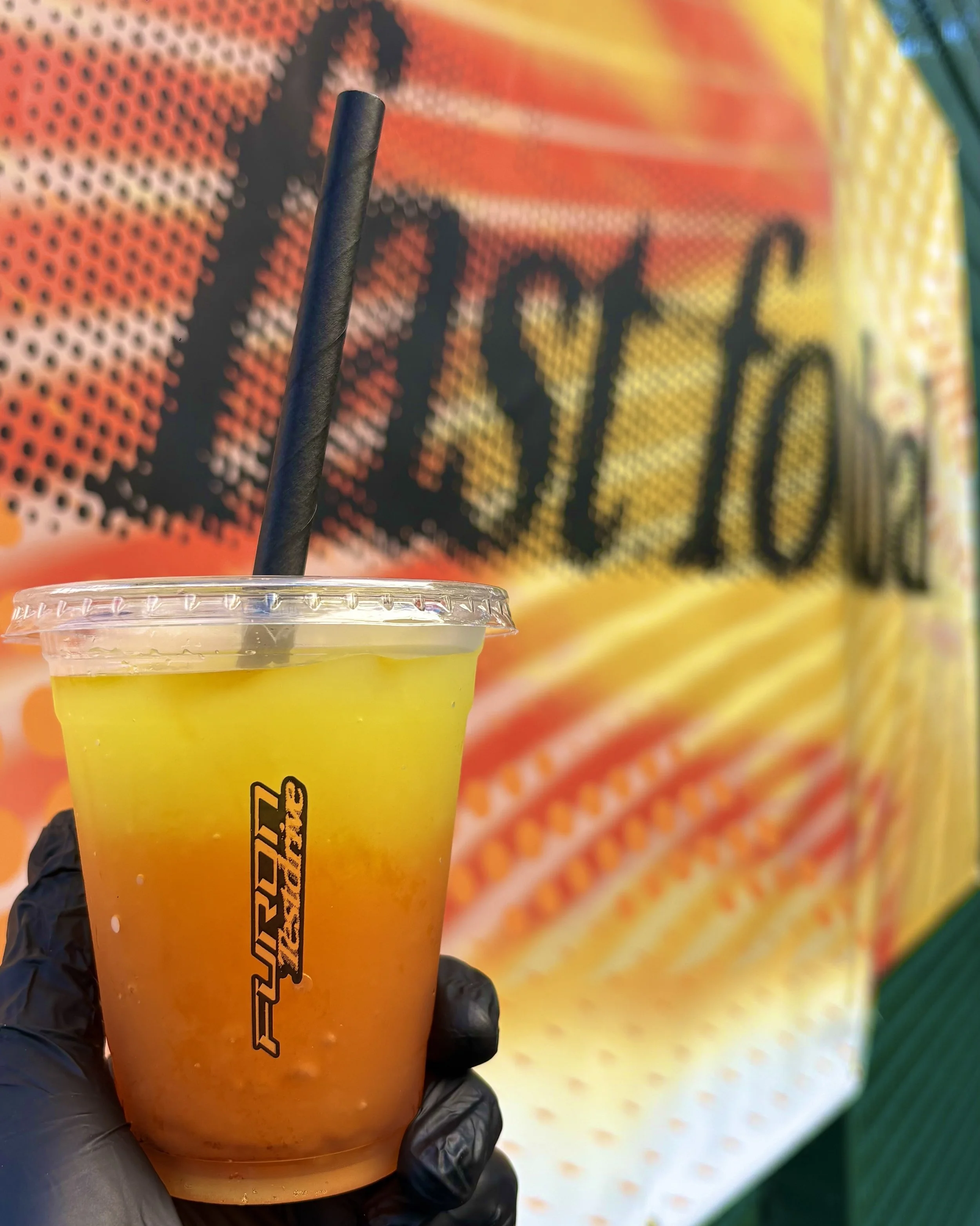 Close-up of a New Balance Furon Test Drive branded cup filled with two-tone pineapple and mango slush, held in front of a wrapped Fast Football signage backdrop.