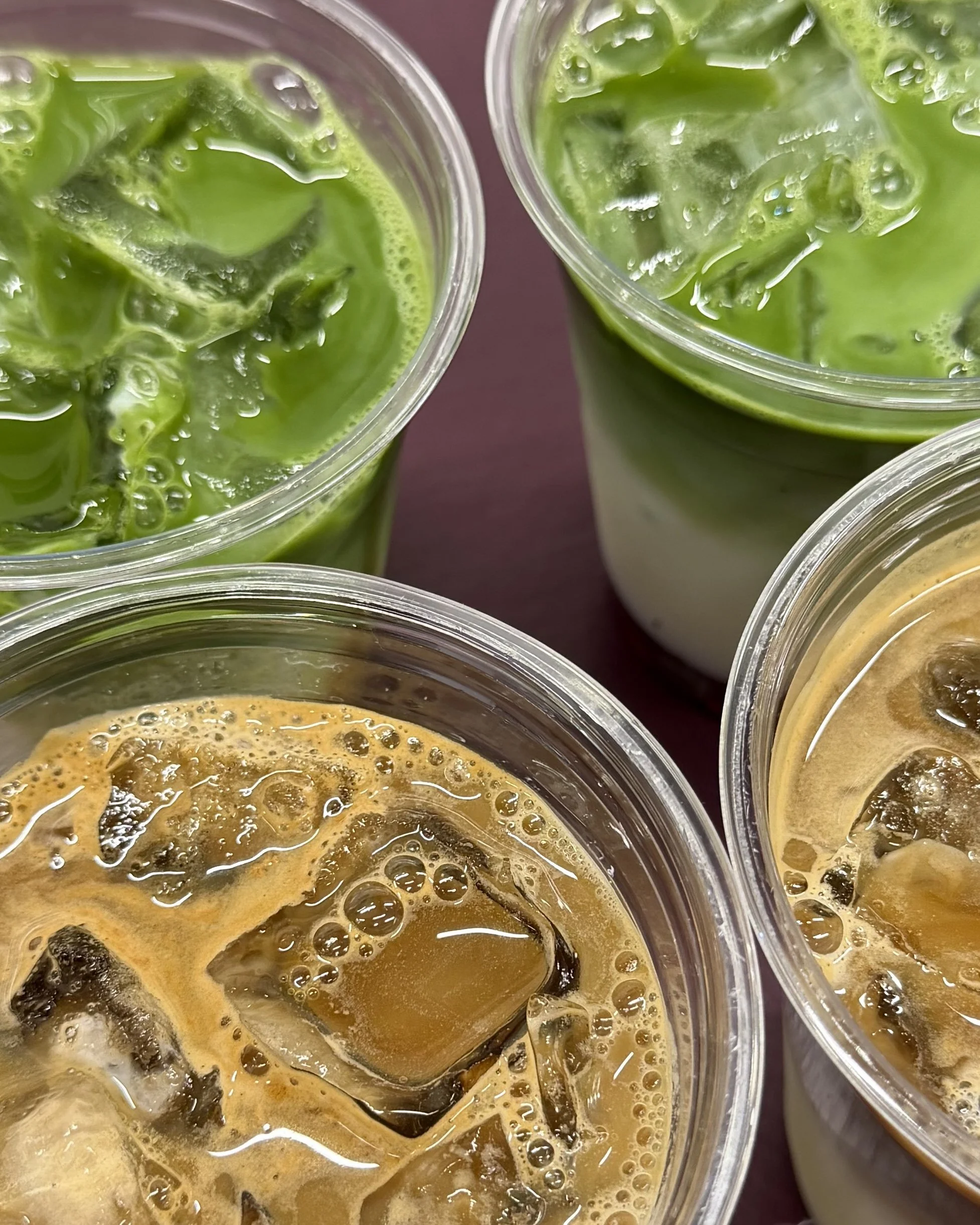 Close-up of four iced drinks served at the Third Floor at The Gilbert coffee and matcha bar, two iced matcha lattes and two iced coffees in clear cups on a burgundy surface.