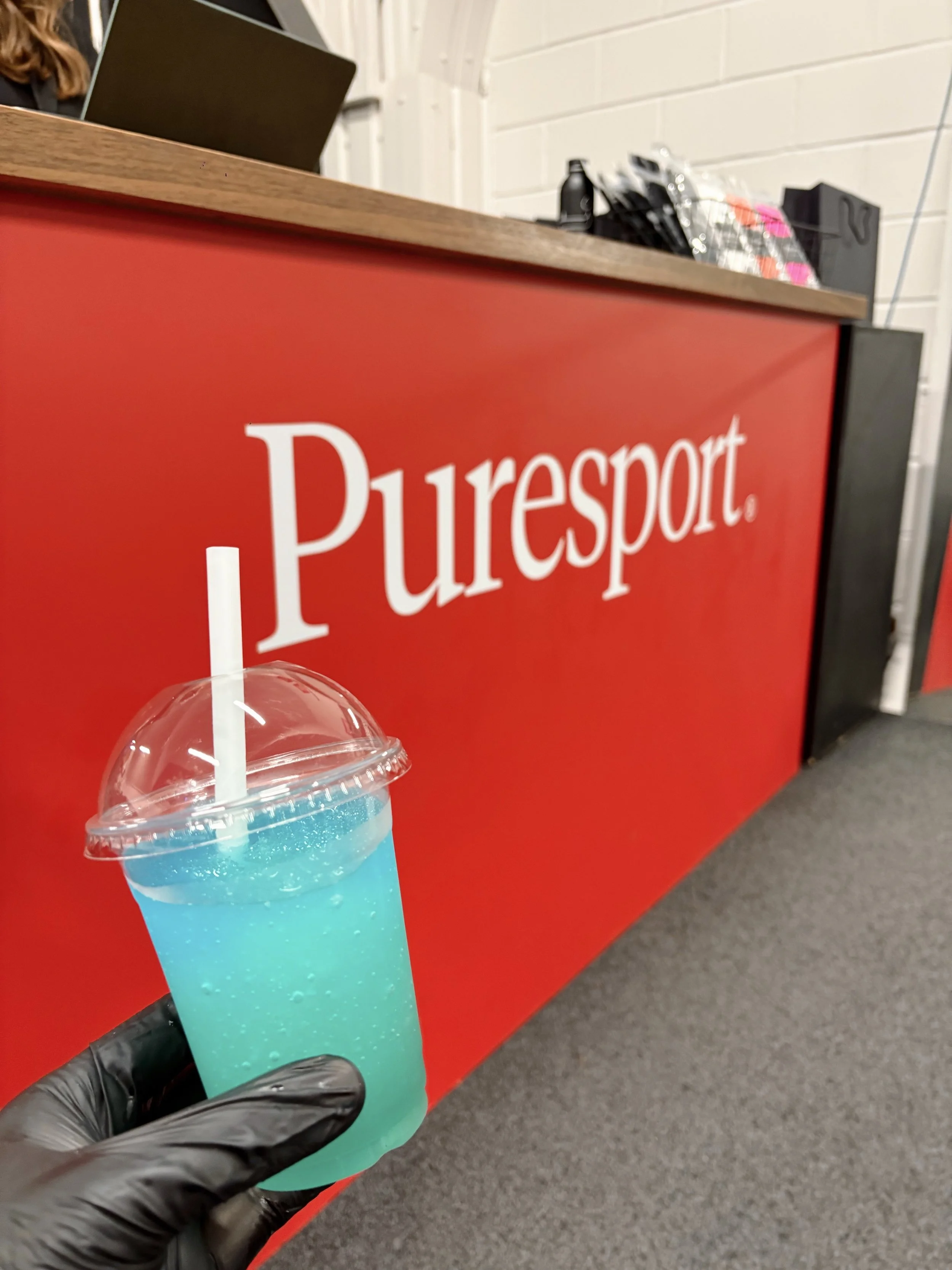 Close-up of a blue slush drink held in front of a red Puresport-branded bar, showing condensation on the cup and a clean, modern event environment.