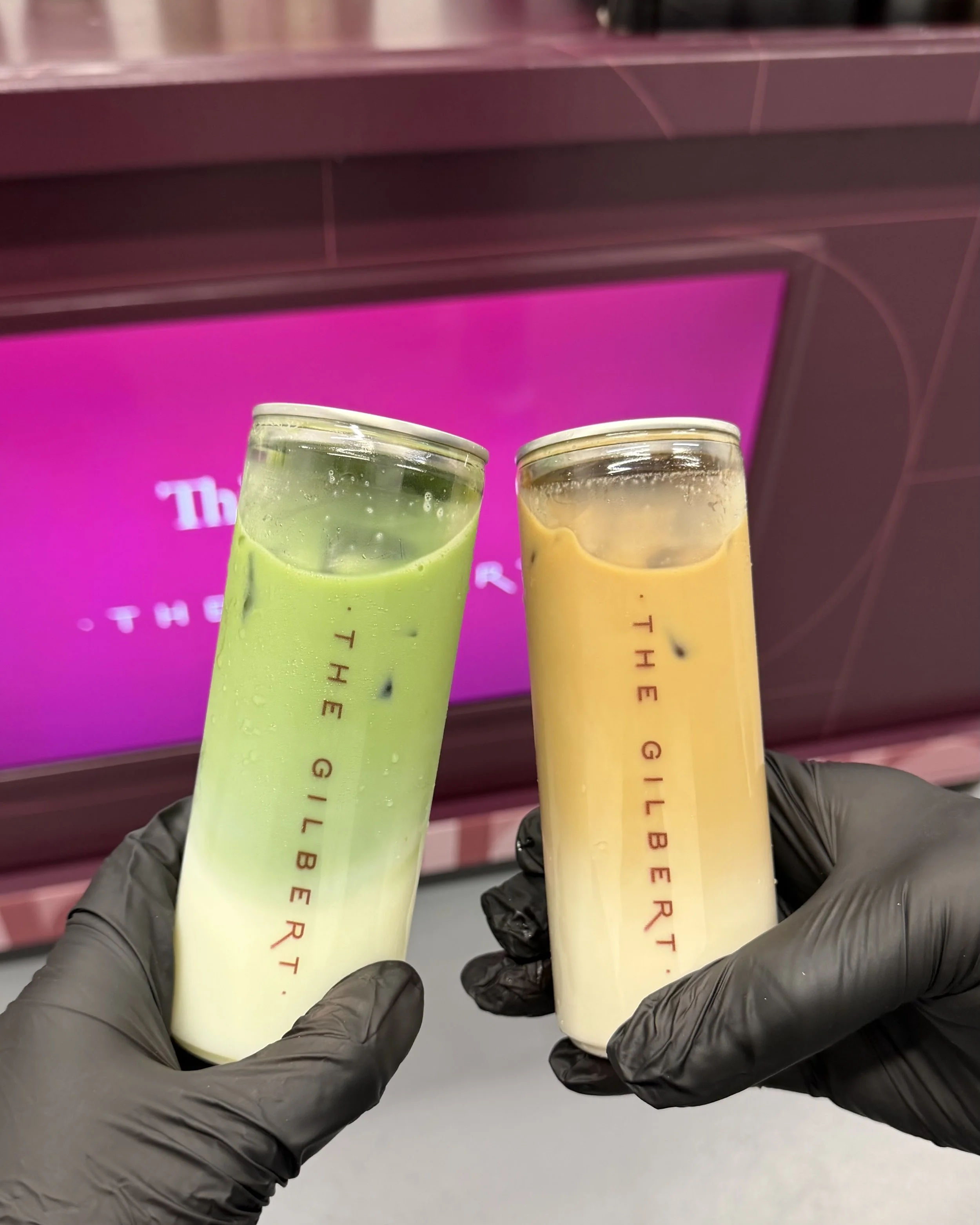 Gloved hands holding two Third Floor at The Gilbert branded cans in a cheers, iced matcha on the left and iced coffee on the right, in front of the burgundy branded bar.