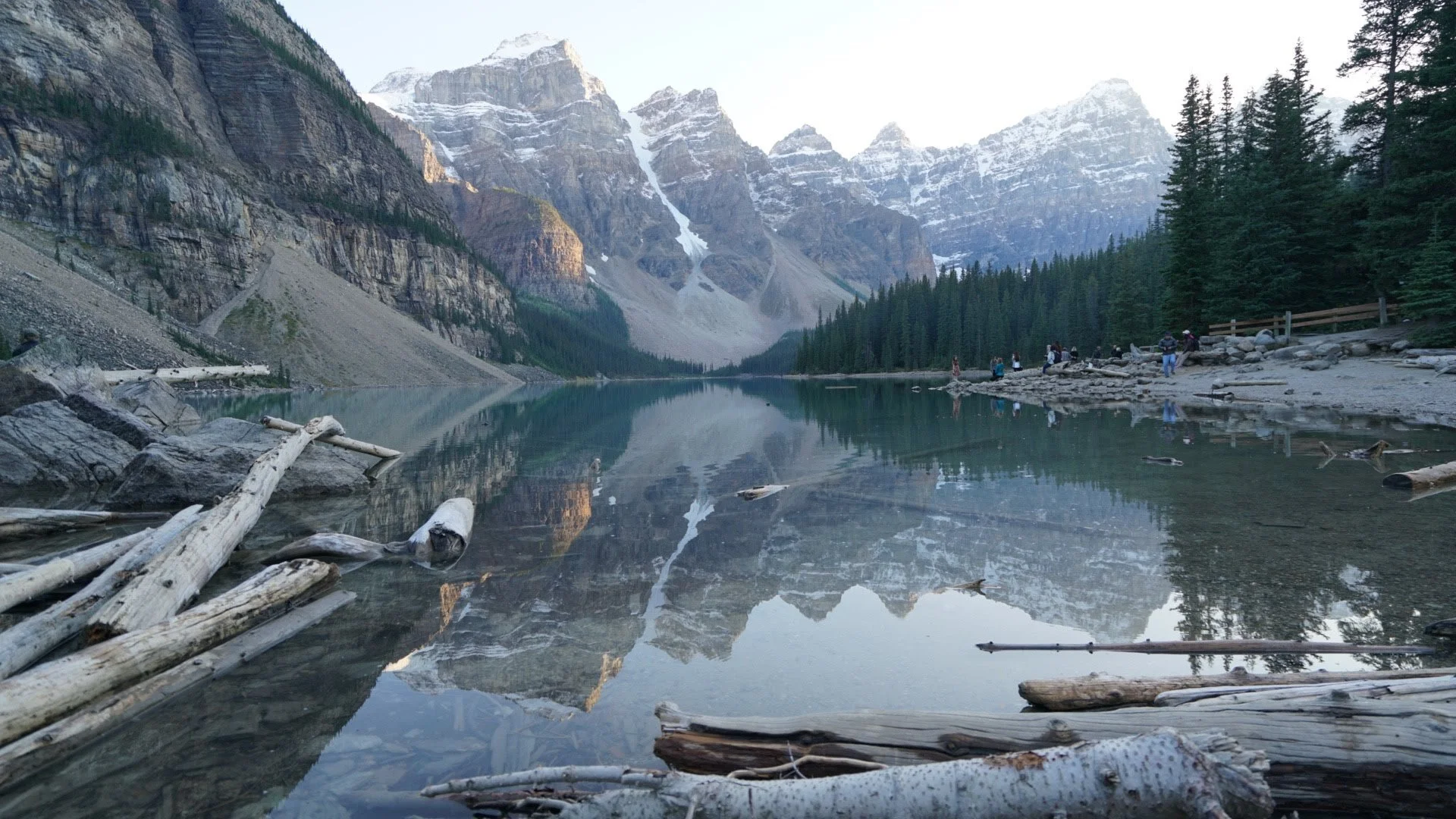 A serene mountain lake with snow-capped peaks and lush pine trees reflected in the water, with logs and rocks along the shoreline and a few hikers on the right.