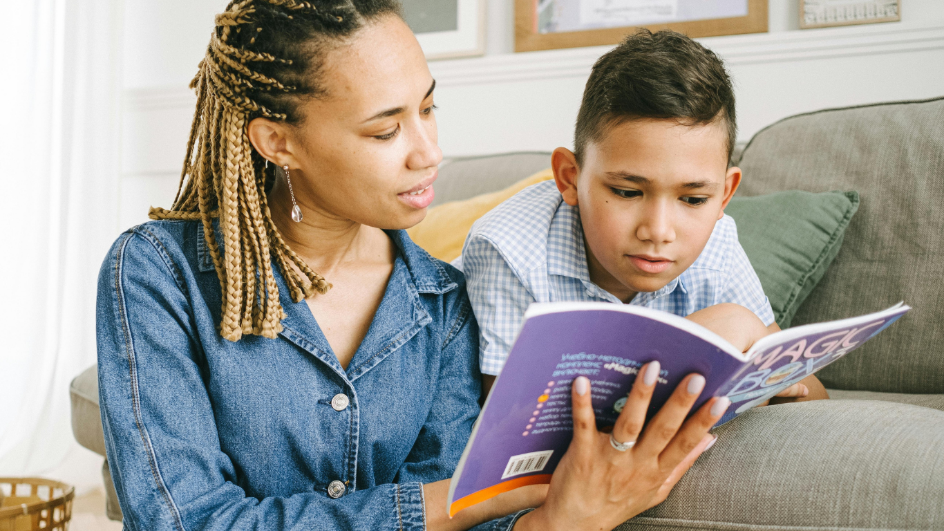 A woman and a young boy sitting on a sofa, reading a book together. The woman has braided hair and is wearing a denim shirt, while the boy has short dark hair and is wearing a light blue checkered shirt. They are focused on the book, which has a purple cover.