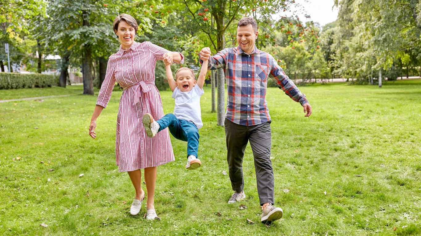 A family of three, a woman, a man, and a young girl, are holding hands and playing together in a park with green grass and trees, smiling and enjoying a sunny day.