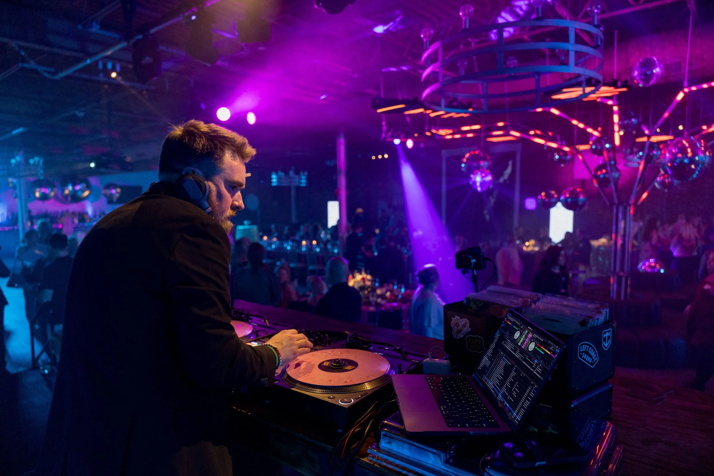 A DJ with a beard and headphones mixing music at a nightclub with purple and pink lighting, surrounded by disco balls and dancing crowd.