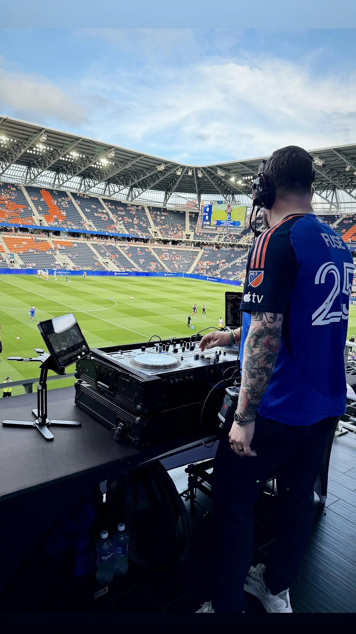 DJ with tattoos wearing headphones and a blue sports jersey playing music at a stadium, with a soccer game and empty seats visible in the background.