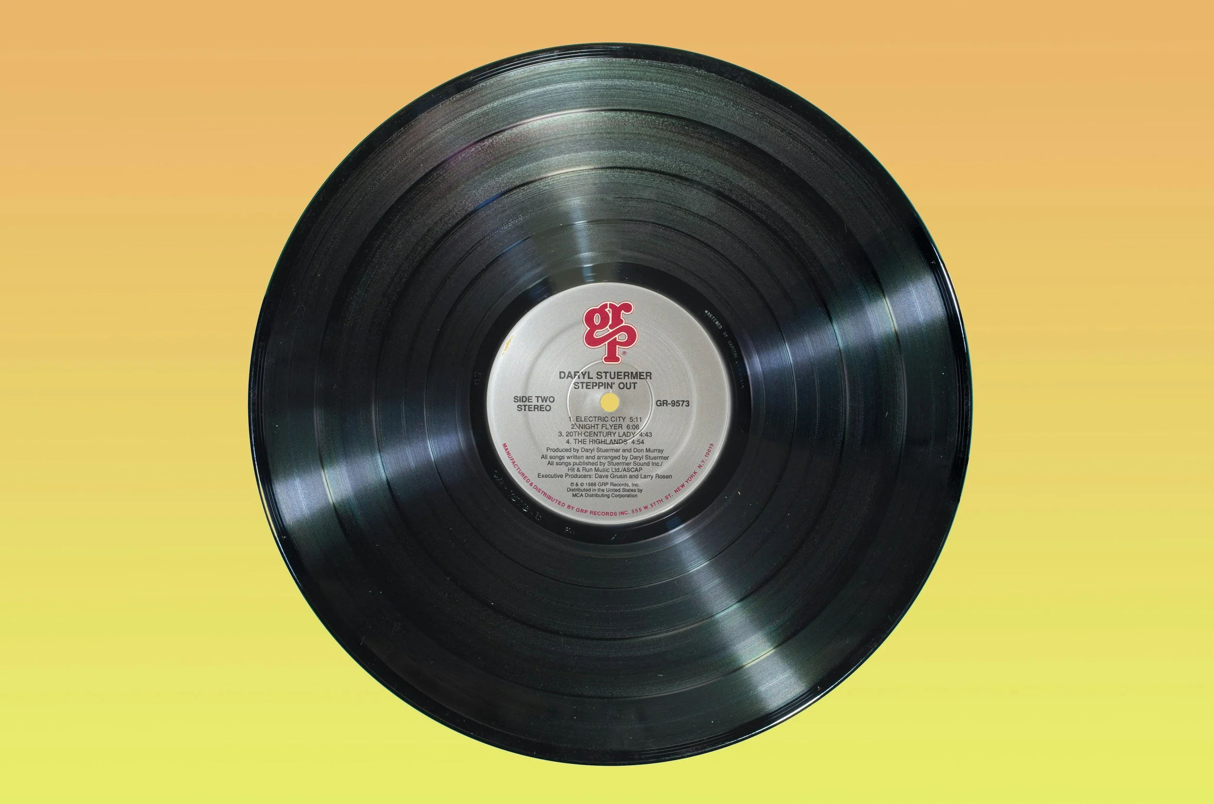 Close-up of a black vinyl record with a white label in the center, displaying red and black text, against a yellow to green gradient background.