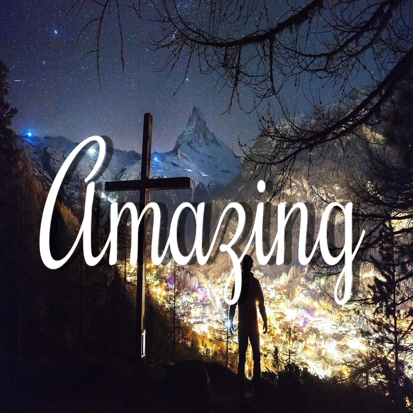 Night view of a mountainous town illuminated with colorful lights, with a person standing in the foreground near a cross, and the starry sky above.