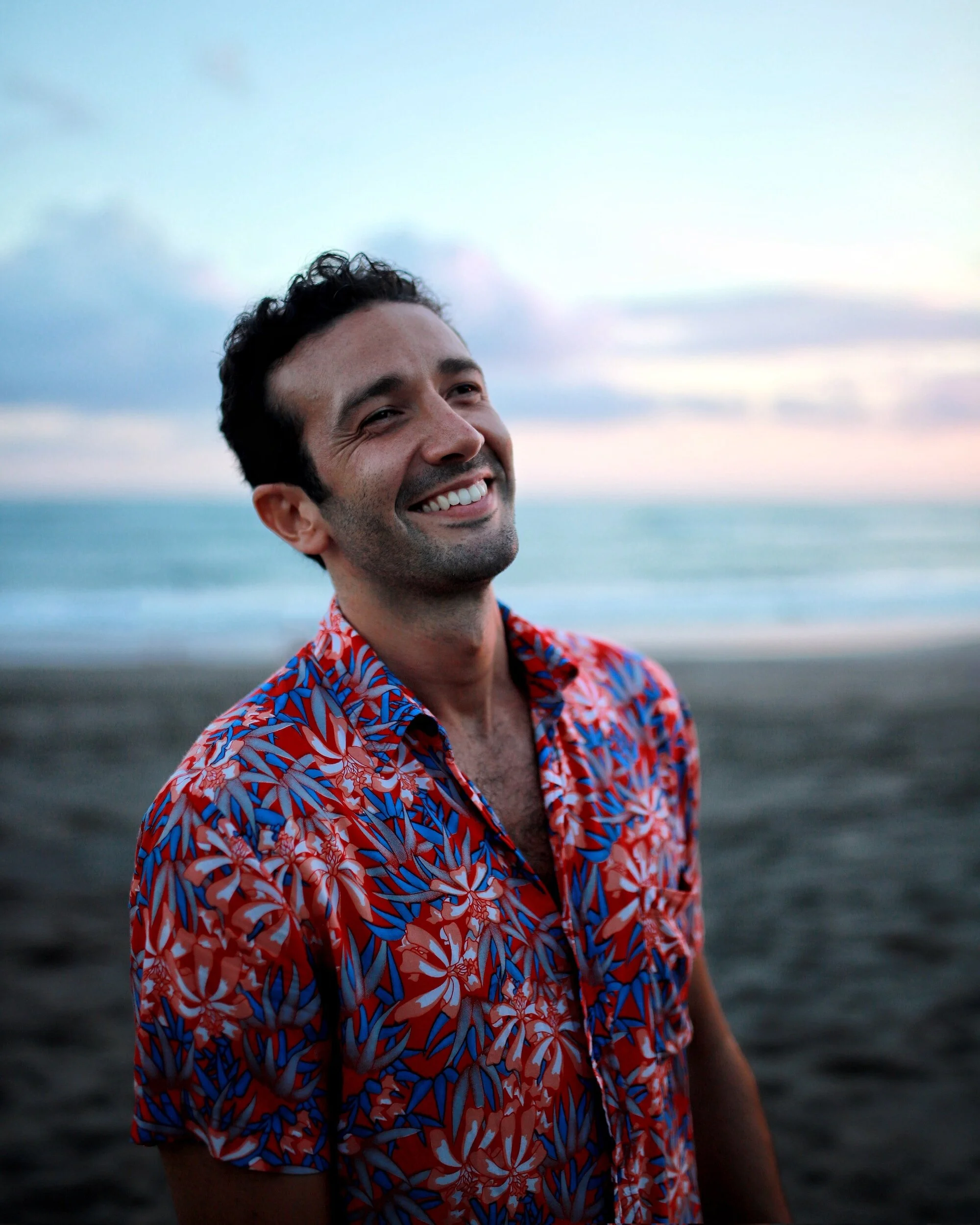 A man standing on a beach during sunset, smiling, wearing a red and blue patterned shirt.