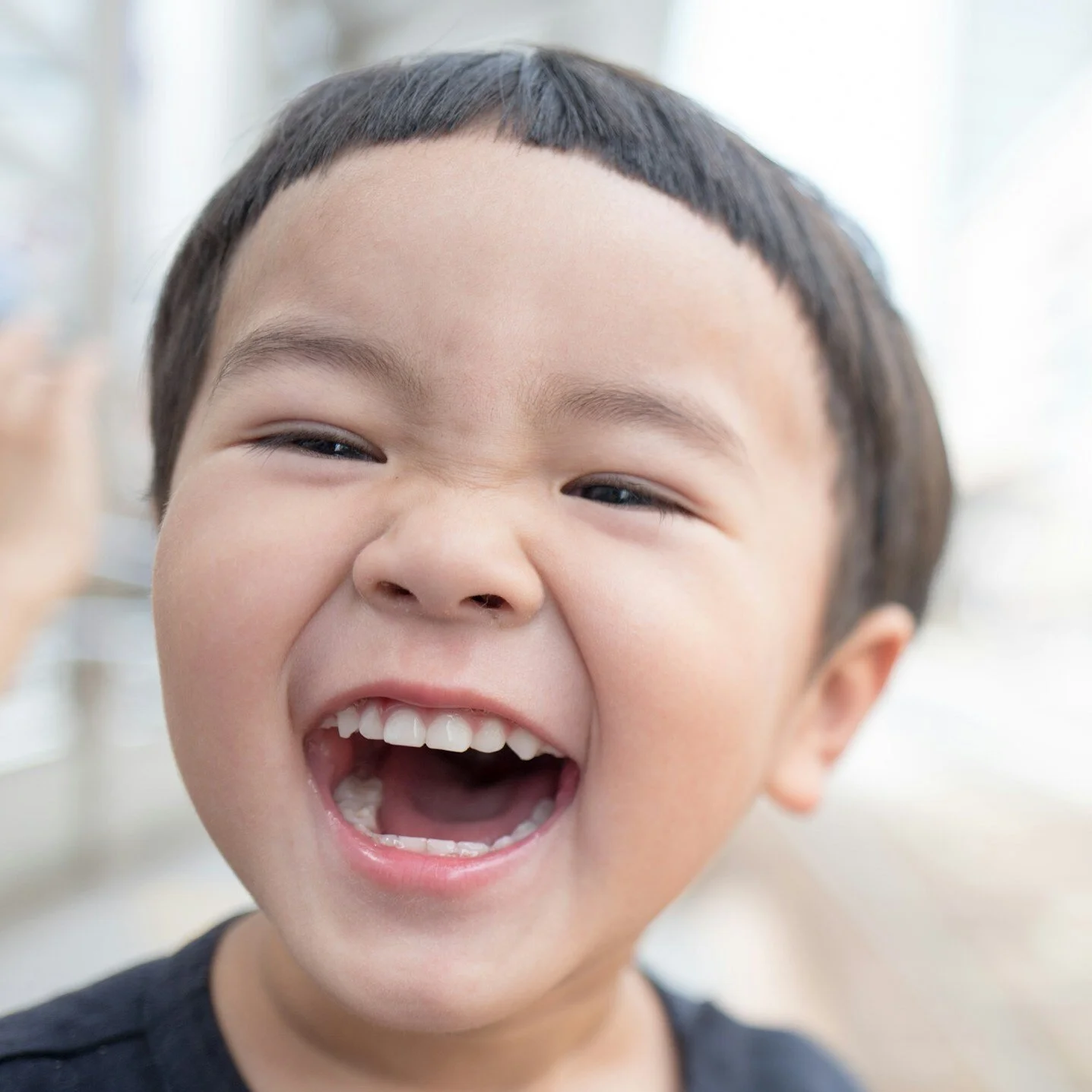 Close-up of a joyful Asian boy laughing with mouth open, showing teeth, in a bright indoor setting.