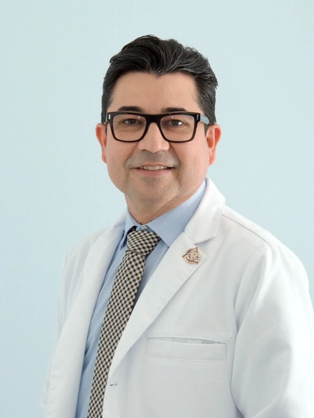 A professional man in a white lab coat, wearing glasses, a dress shirt, a patterned tie, and an insignia pin on the lapel, smiling at the camera.