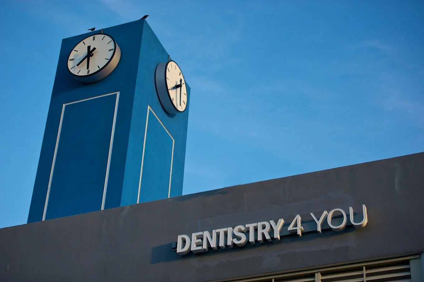A blue clock tower with two clocks showing 11:25, against a blue sky, located above a building with a sign that reads 'DENTISTRY 4 YOU'.