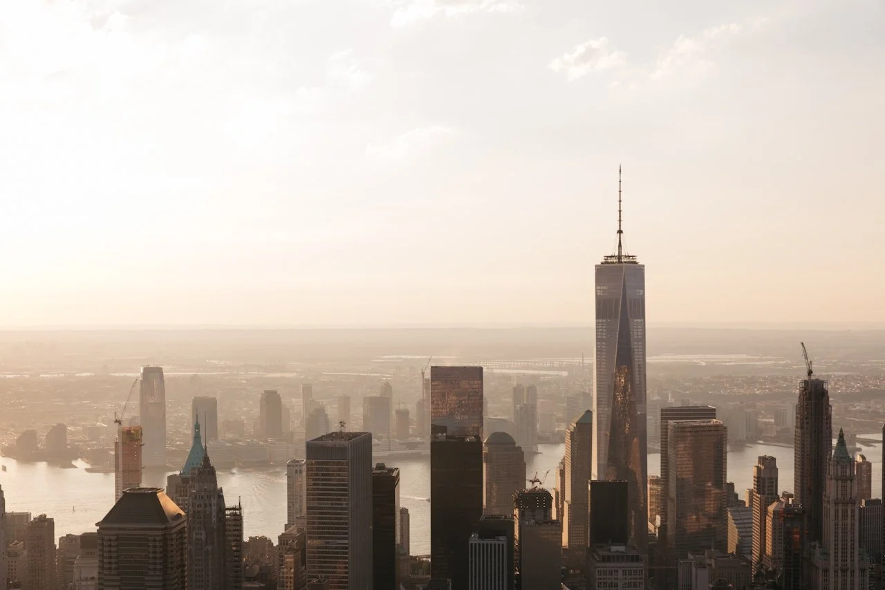 Wide view of a city skyline at golden hour, featuring tall skyscrapers and a river in the distance under a hazy sky.