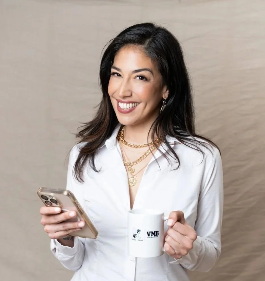 Vanessa is smiling, holding a smartphone in one hand and a white coffee mug in the other, wearing a white shirt and gold jewelry, standing against a beige background.