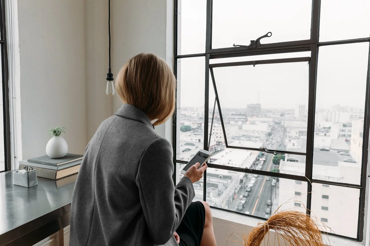 Businesswoman in a gray coat sitting by a large industrial-style window, holding a smartphone and overlooking a cityscape on a cloudy day