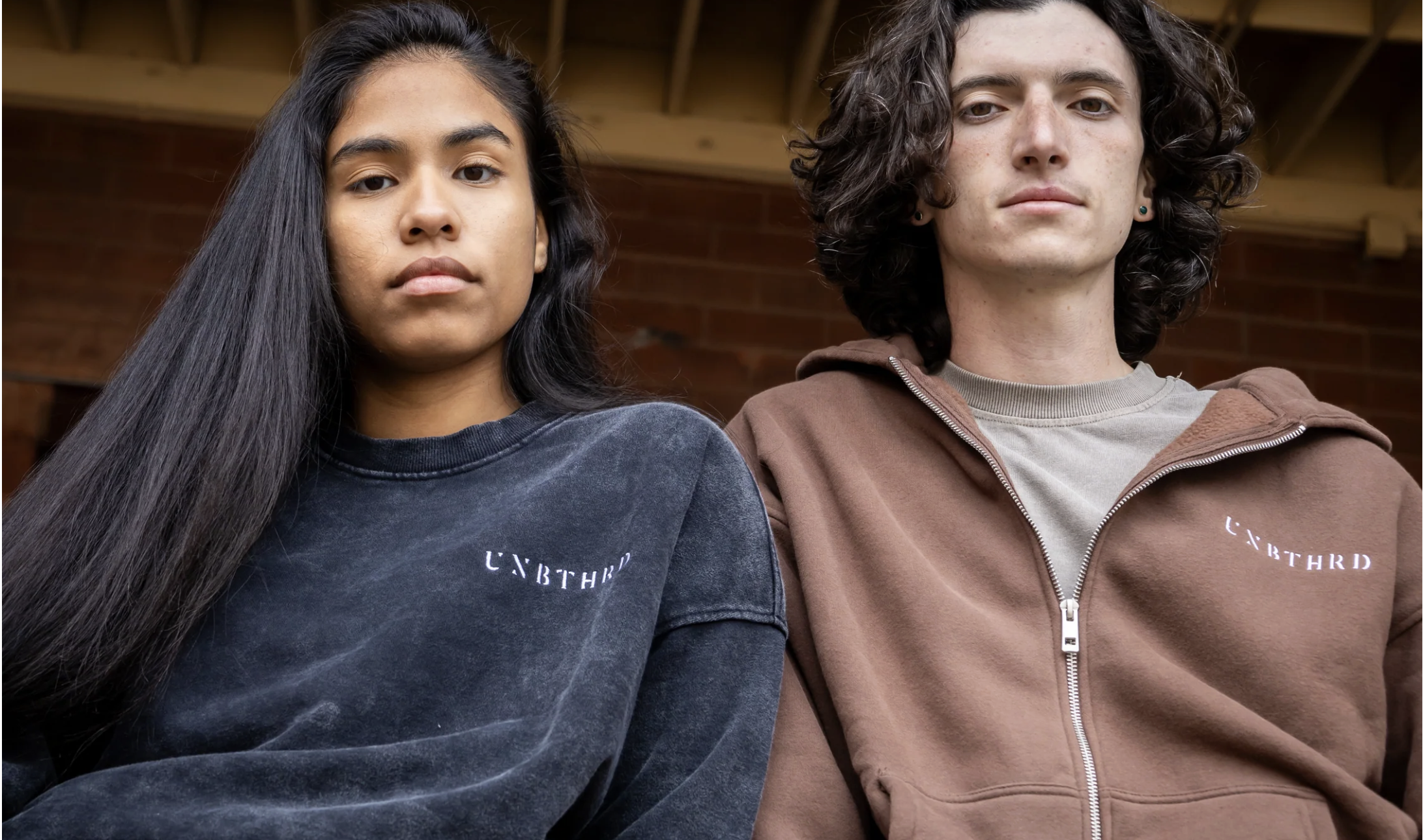Two models standing side by side outdoors, wearing casual sweatshirts and hoodies with a minimalist logo, photographed from a slightly low angle.