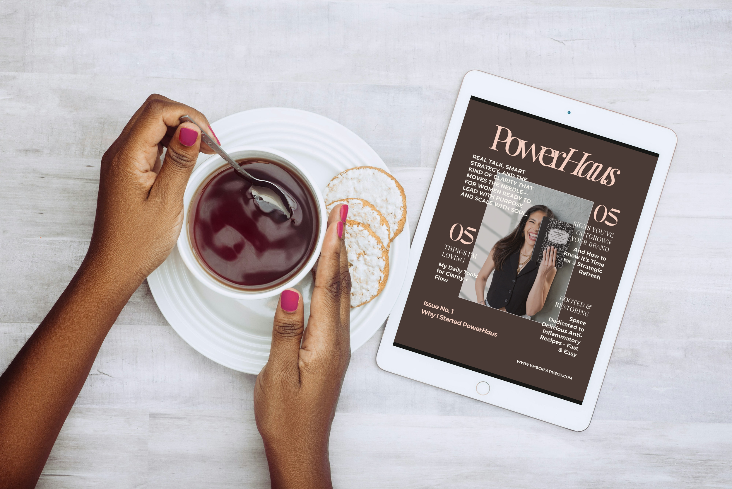 A person's hands with pink nails holding a spoon in a cup of dark tea on a white plate with three sugar cookies, next to an iPad displaying a magazine cover titled 'PowerHaus' on a light-colored wooden surface.