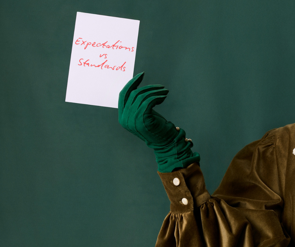Gloved hand holding a small card with handwritten text reading “Expectations vs Standards” against a solid green background.