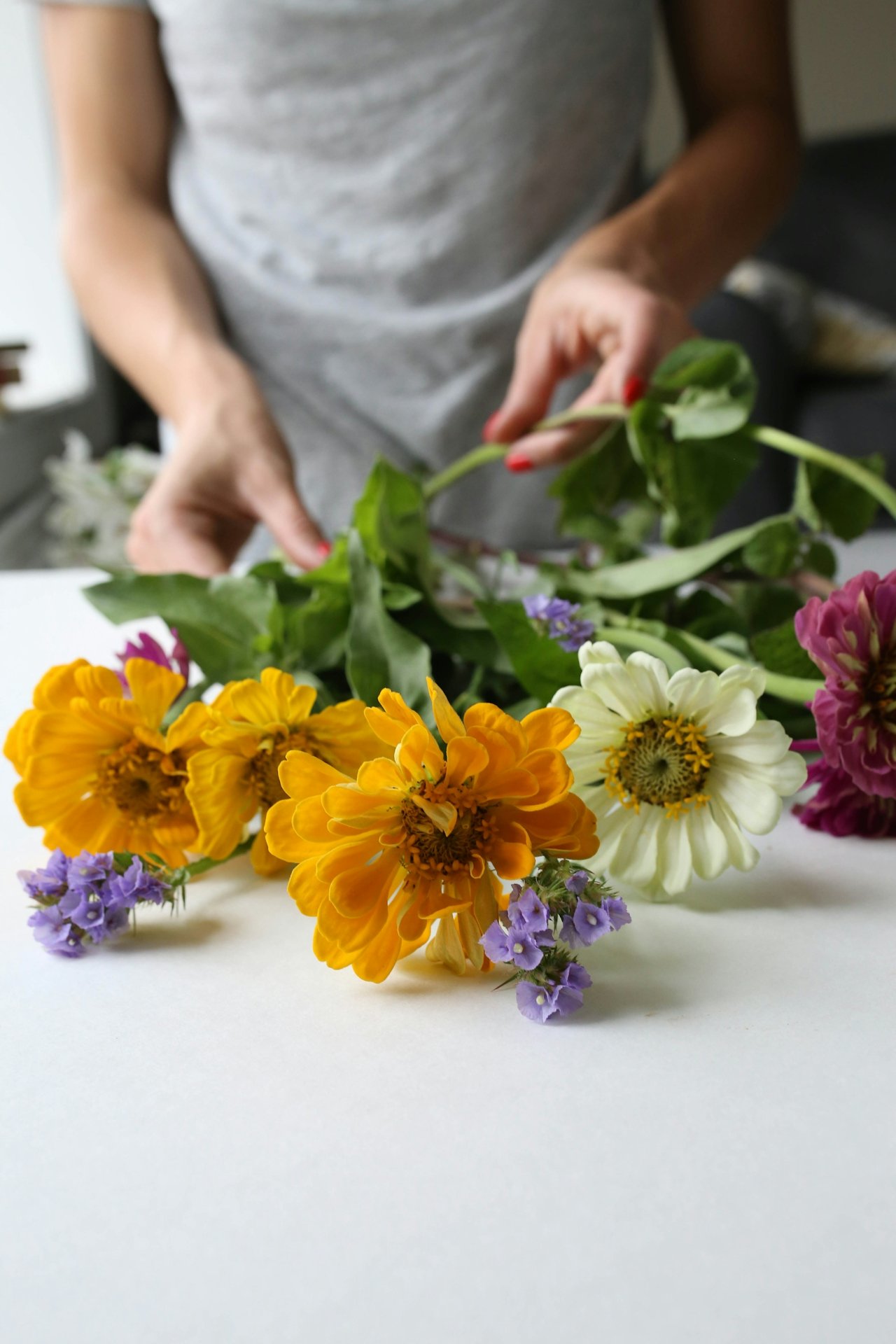 Close-up of a person arranging a colorful bouquet with orange, white, purple, and magenta flowers on a white surface