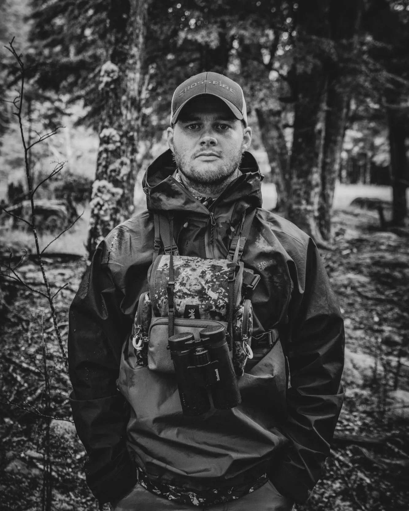A black-and-white photo of a man in outdoor gear standing in a forest. He is wearing a cap, a jacket, and has binoculars hanging from his neck.
