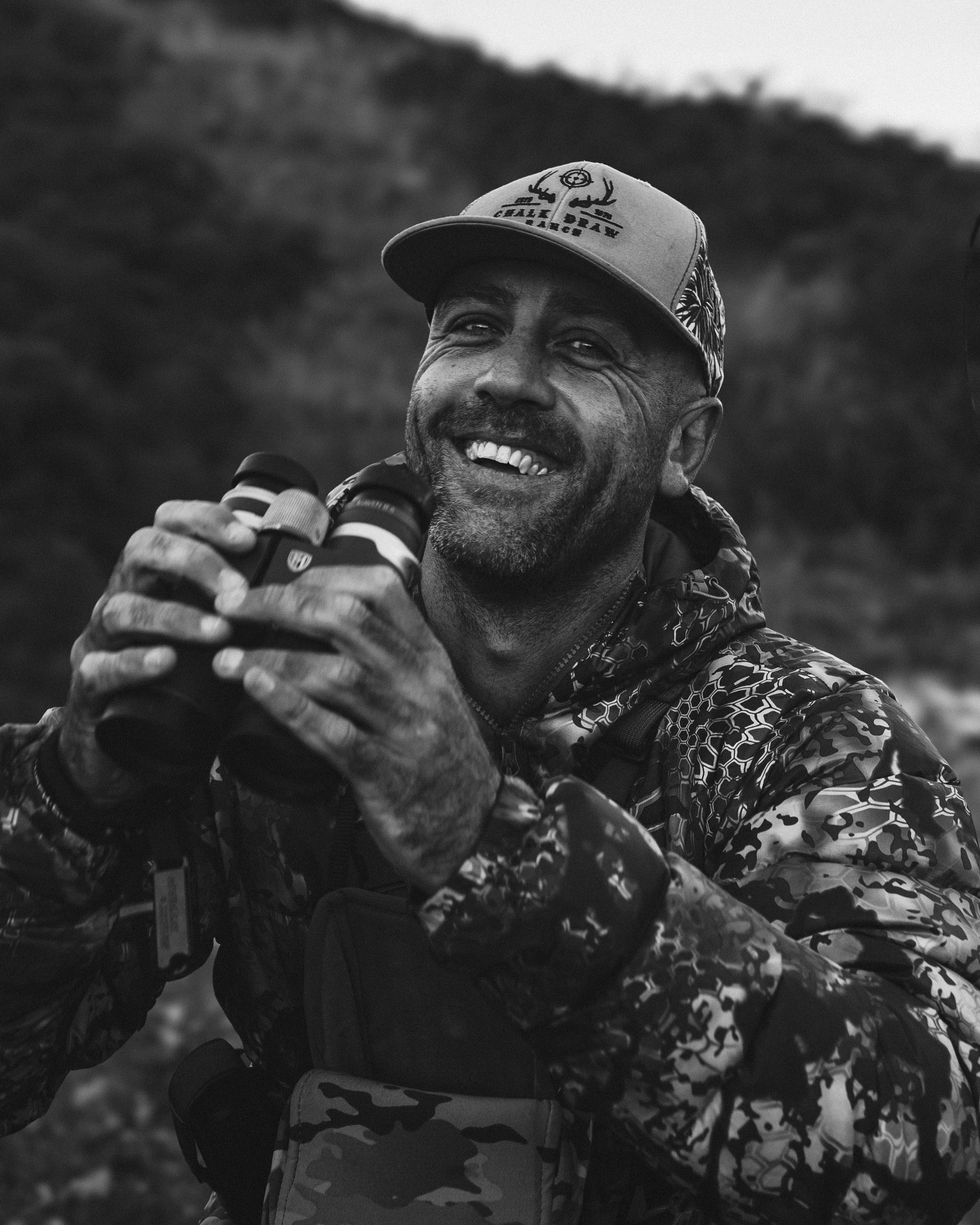 Flow State Productions Founder Wade Kelly, holding binoculars, wearing a camouflage jacket, a cap with deer antlers and text, outdoors with a blurred hillside background.