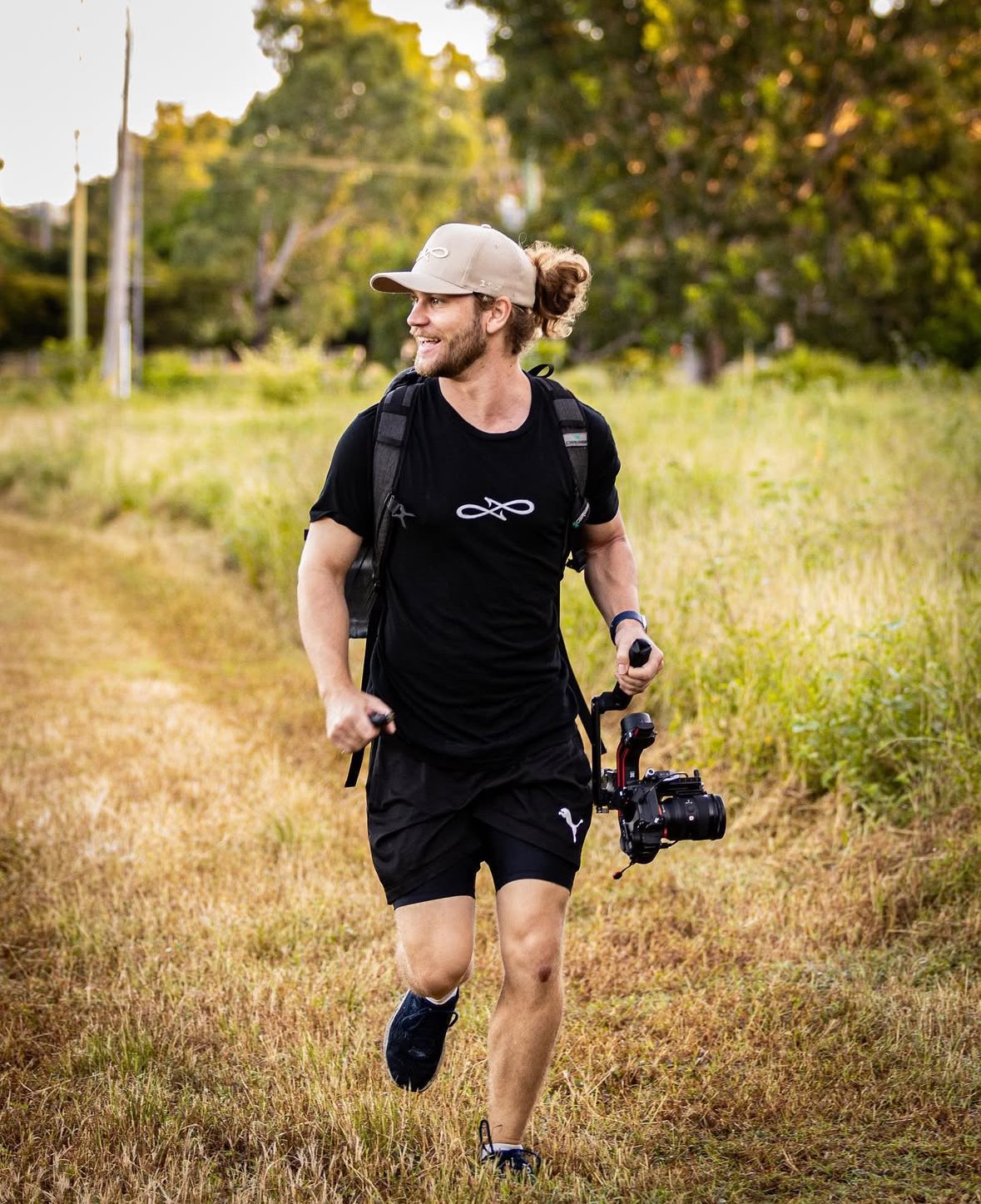 A young man with long hair tied in a bun, wearing a beige cap and black athletic clothes, running outdoors on a grassy trail with a camera in hand and a backpack, smiling. Flow State Productions Outdoor hinting Photography & videography