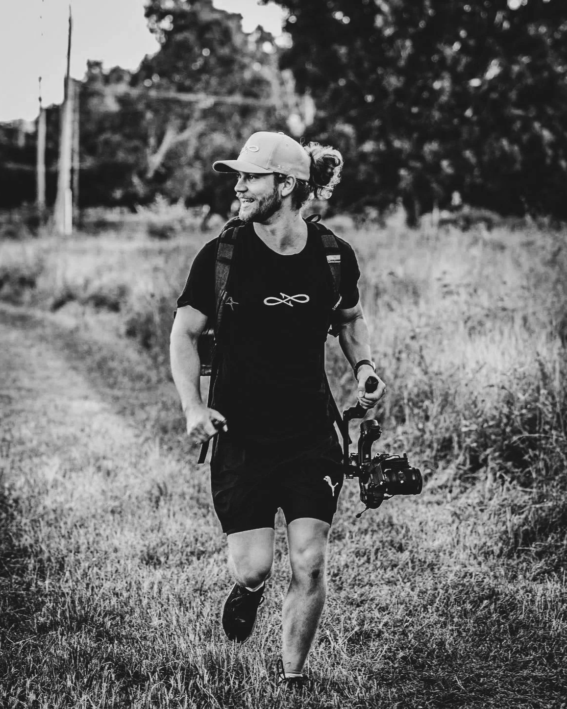 A man with a beard and long hair tied back, wearing a cap, running outdoors on grass with a camera hanging from his waist, and carrying a backpack, smiling and looking to the side.