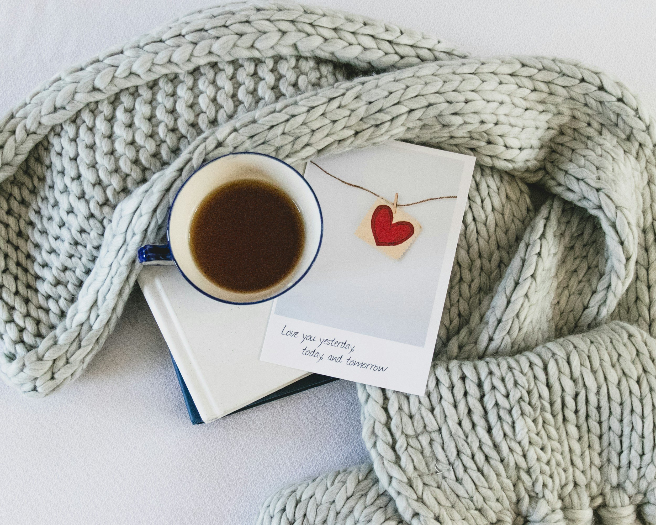 A white knitted blanket draped over a white surface, with a white book, a cup of coffee, and a photo with a red heart on string.