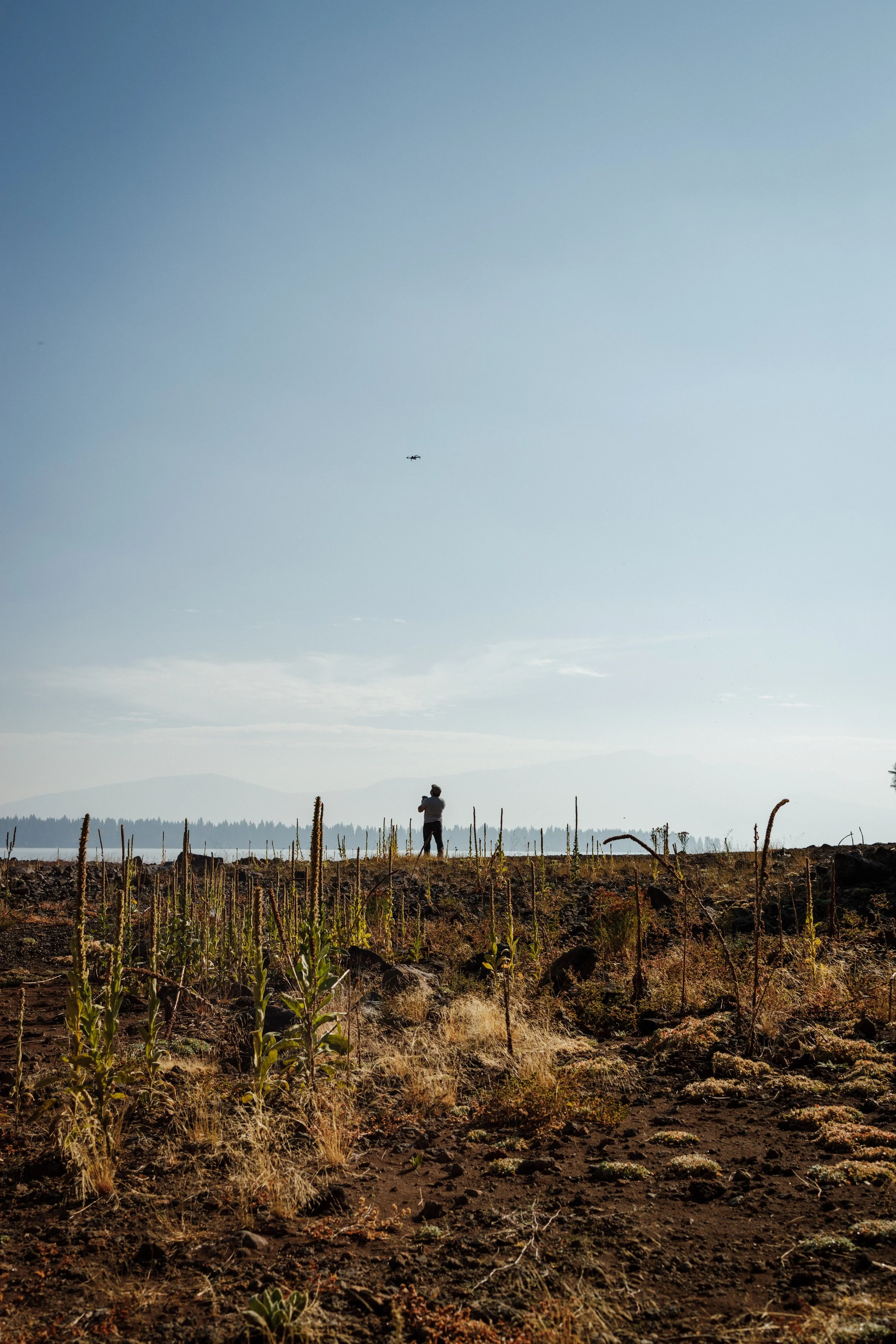 A person flying a drone in a dry, rocky field with sparse vegetation and a clear sky.