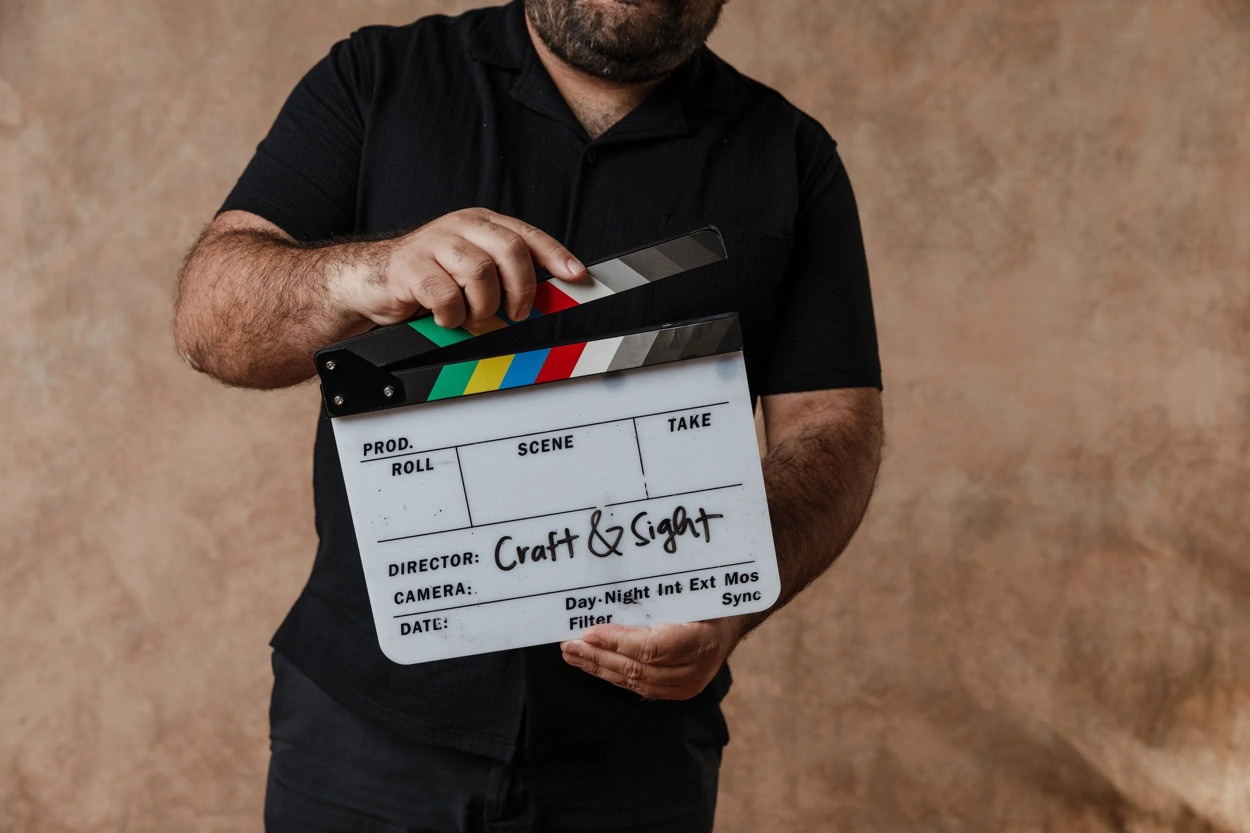 Man in black shirt holding a director's clapboard with 'Craft & Sight' written on it, standing against a brown textured background.