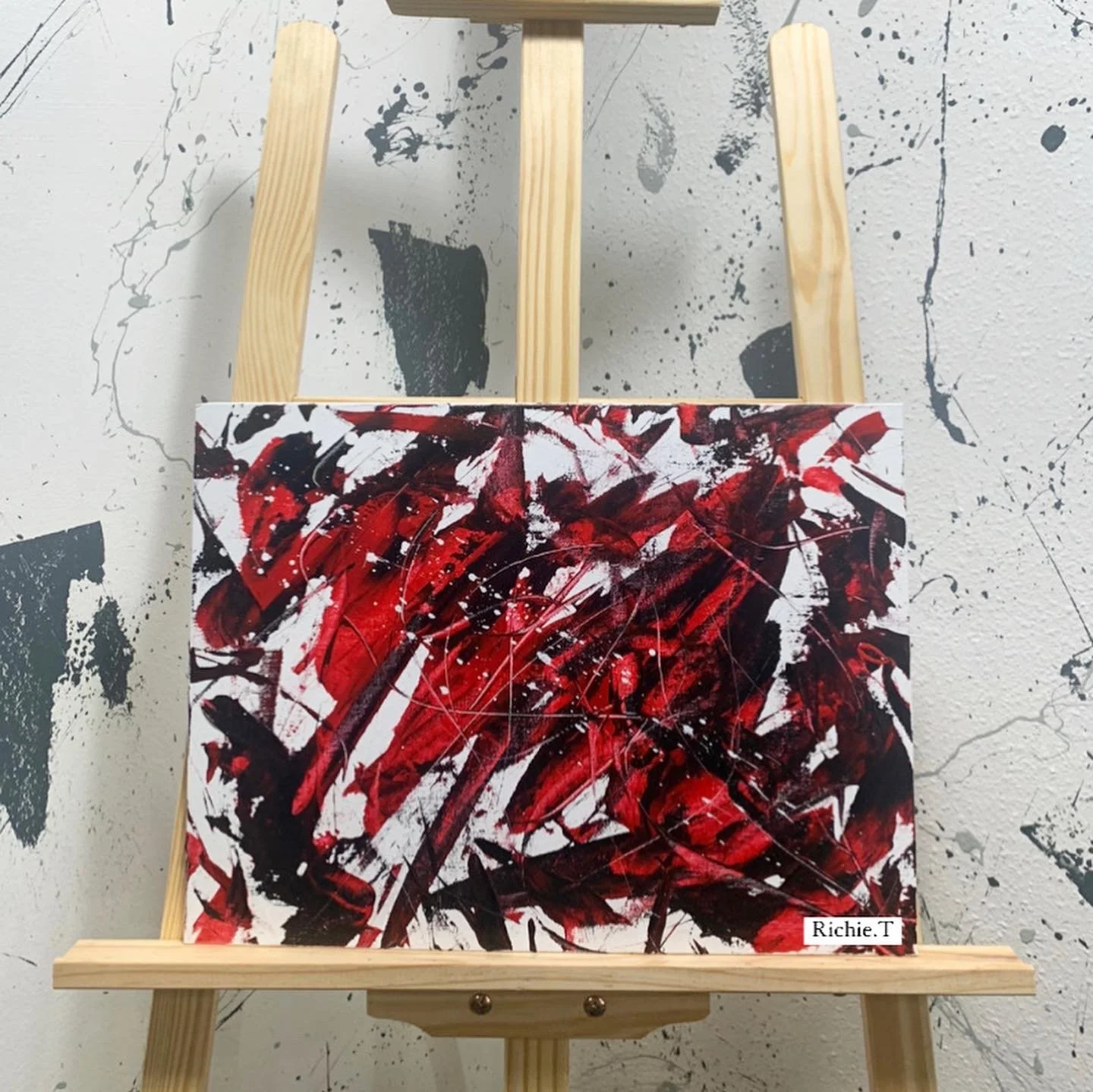 Abstract painting with red, black, and white splashes on a canvas placed on a wooden easel against a wall with splatter paint background.