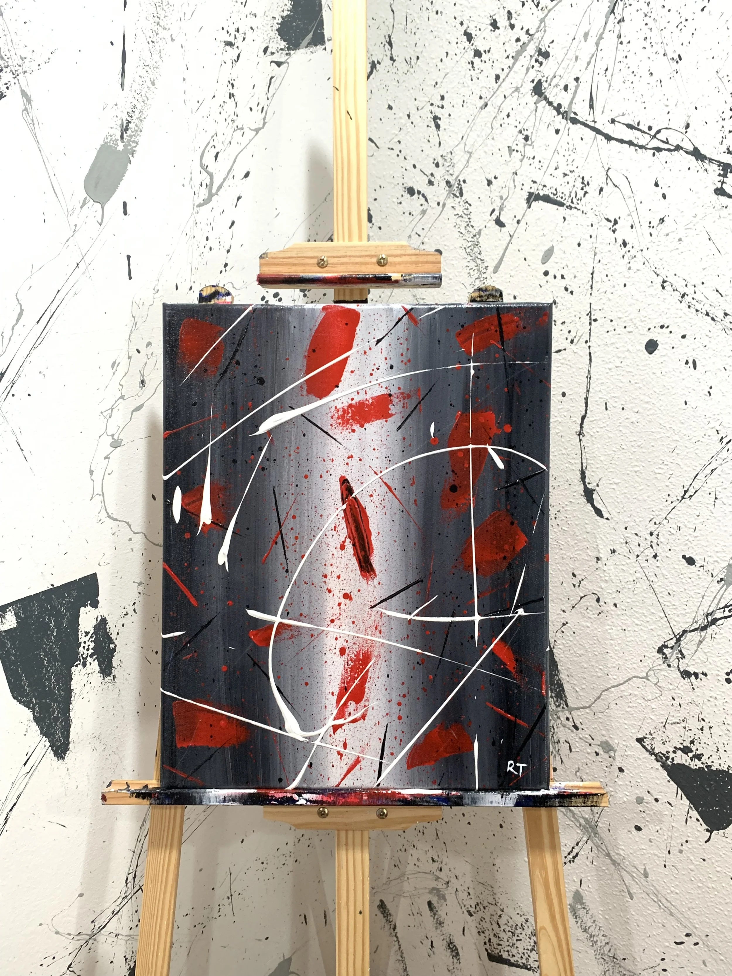 Abstract painting on a canvas featuring splashes and streaks of red, white, black, and gray, mounted on a wooden easel against a splattered black and white wall.