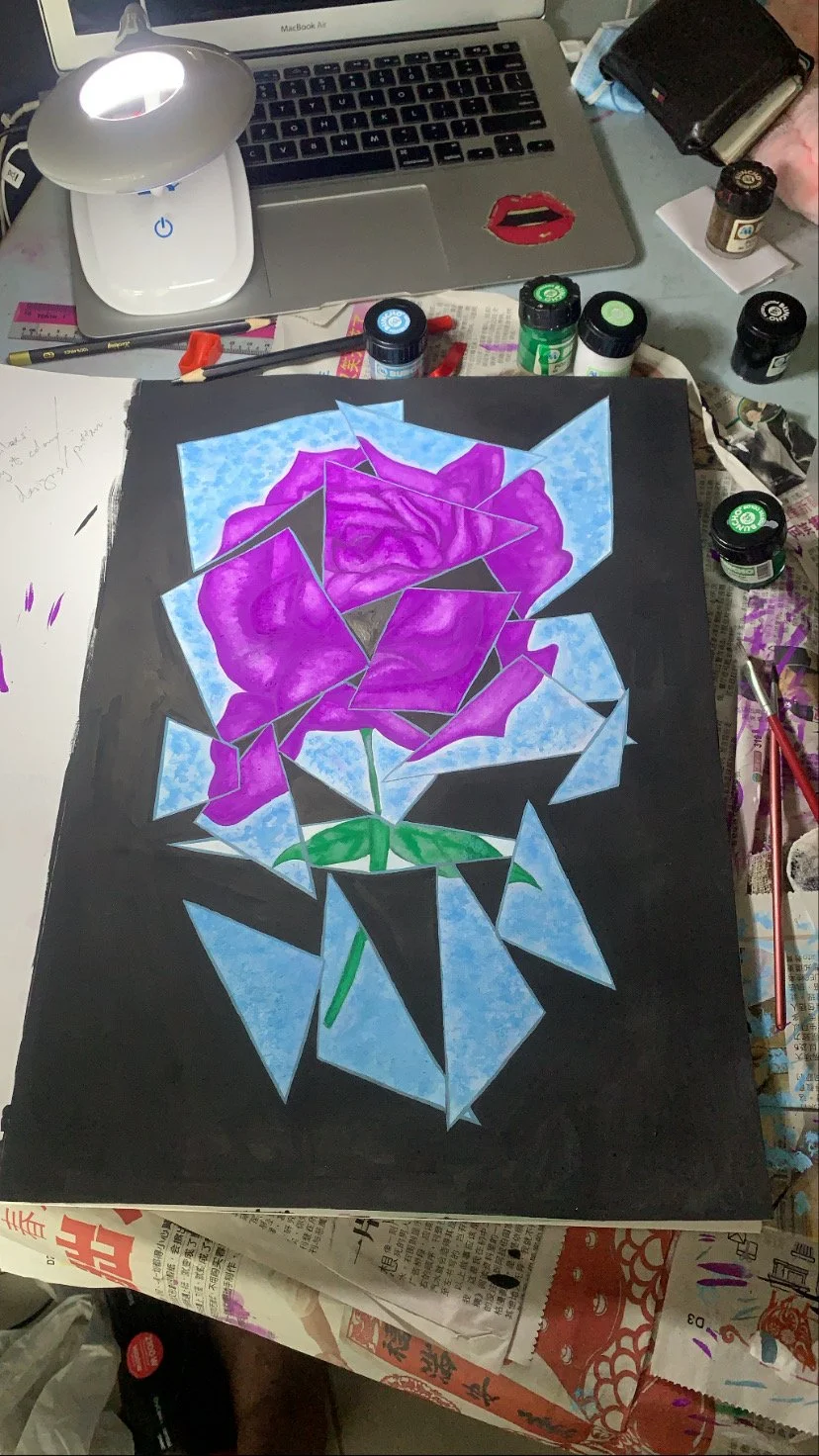 A geometric painting of a purple rose with green leaves on a black background. The painting is surrounded by art supplies, a laptop, and a desk lamp.