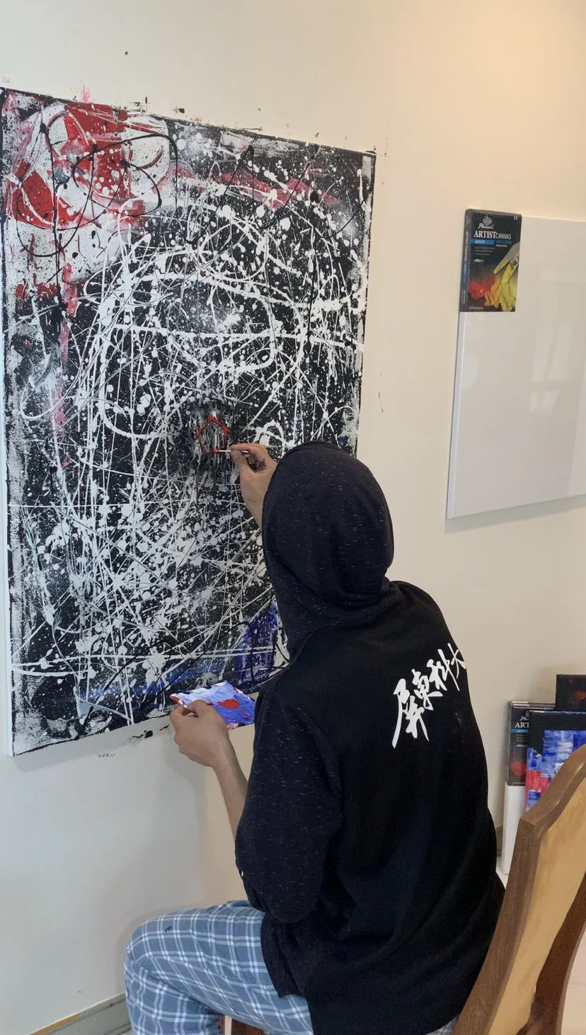 A person dressed in black with Asian writing on the back is painting abstractly with red and white paint on a black canvas with a splattered and scribbled pattern.