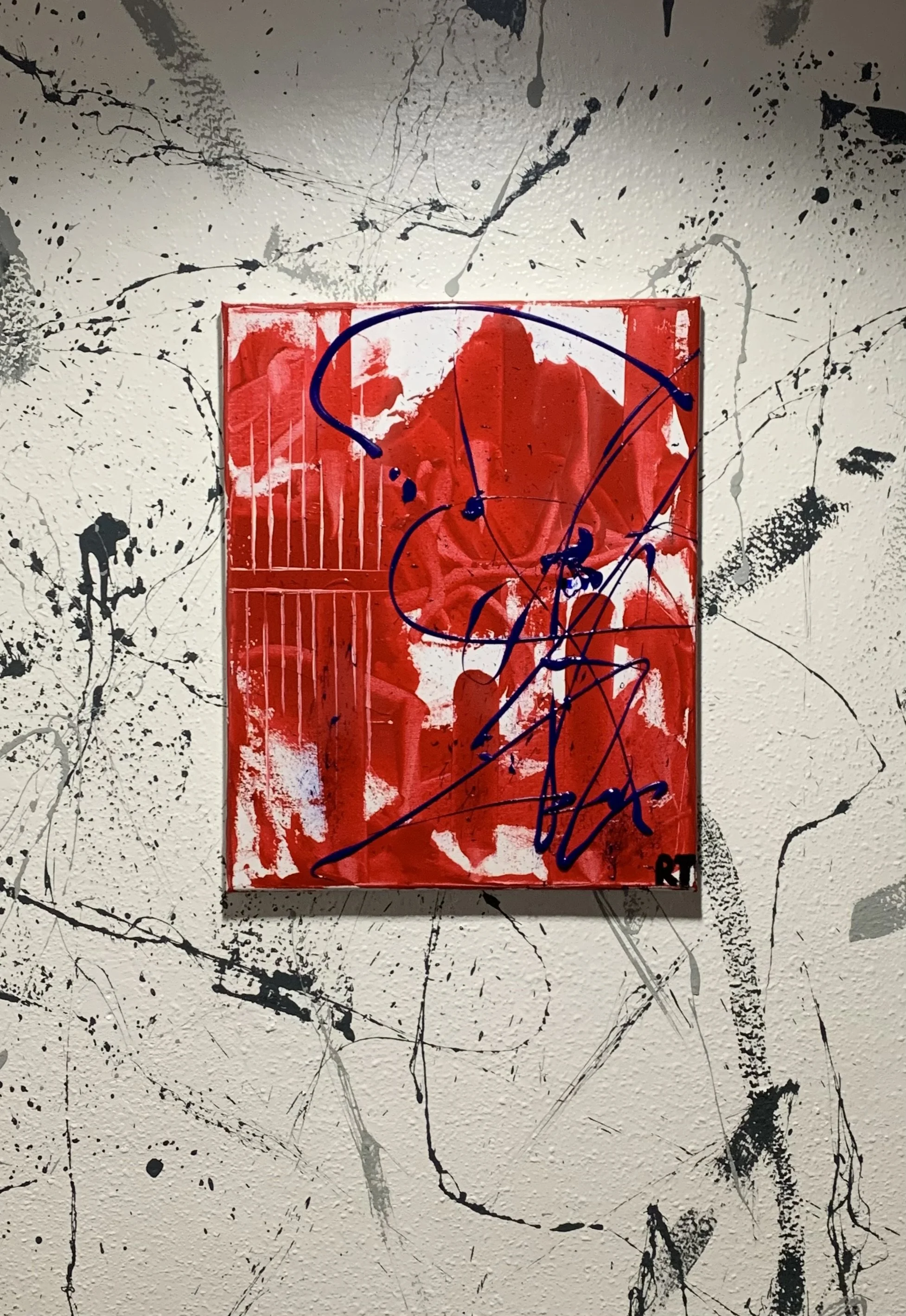 Abstract graffiti art on a textured wall featuring a small red canvas with black and blue spray paint, drips, and scribbles.