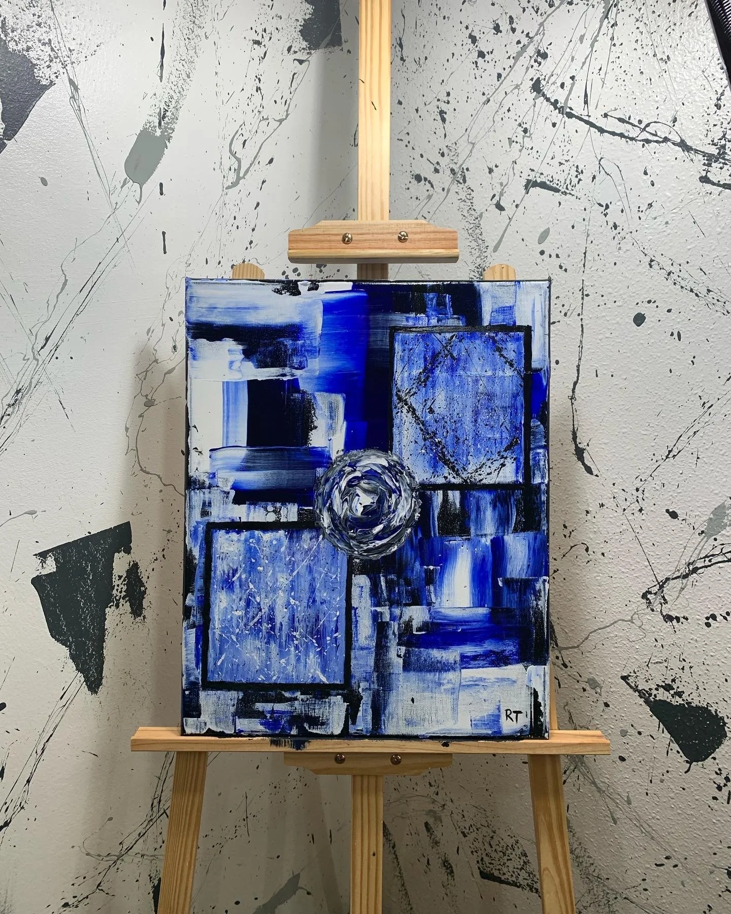 Abstract painting with shades of blue, black, and white on a small canvas, displayed on a wooden easel against a splattered black and white background wall.