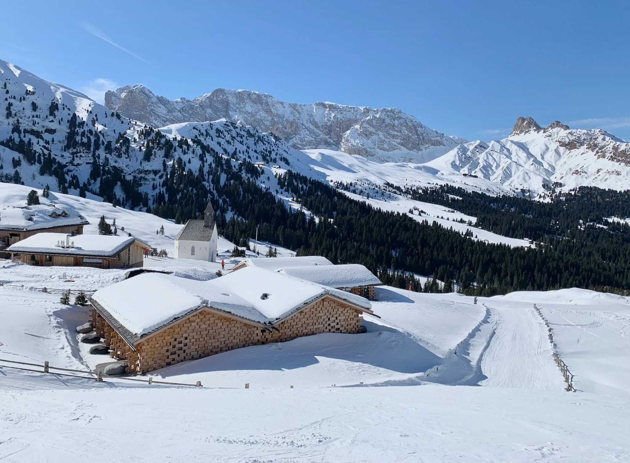 Snow-covered wooden buildings and trees in the mountain landscape of the Dolomites of the Alpe di Siusi, in Trentino-Alto Adige, Italy, with a small white church and ski tracks.