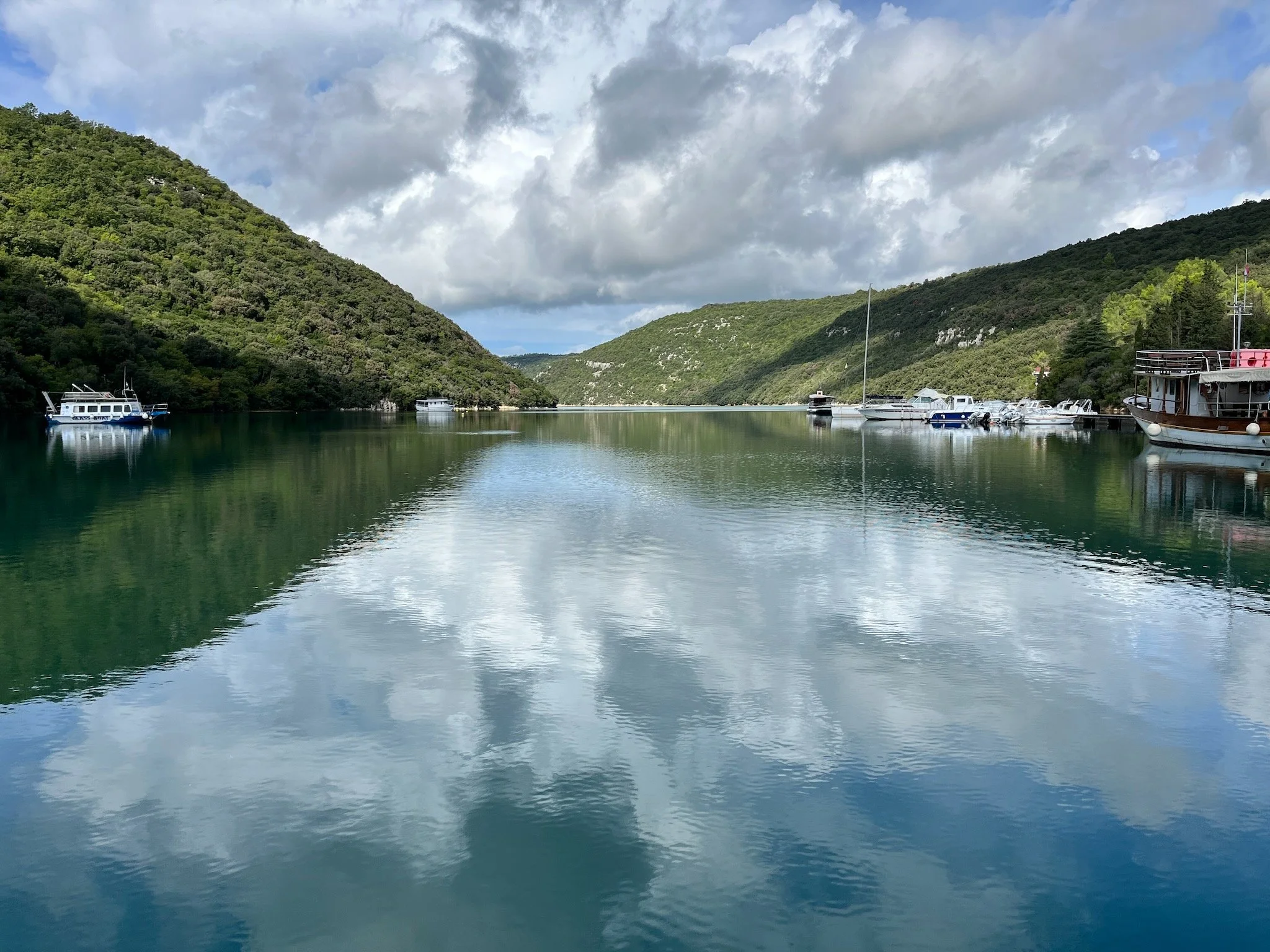 The calm waters of the Adriatic Sea in the Lim fjord, in Istria, Croatia, surrounded by green, forested hills, with several boats anchored along the shoreline. The sky is partly cloudy, and the water reflects the clouds and hills.