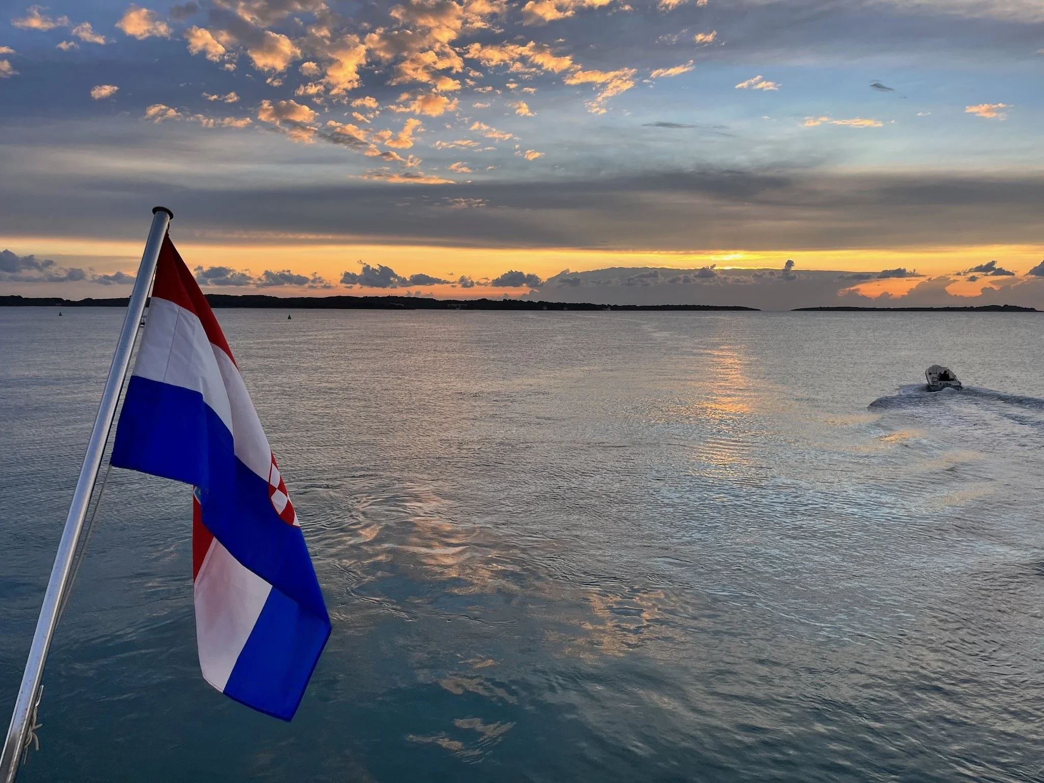 A boat on the calm water of the Adriatic Sea at sunset with a Croatian flag in the foreground and a distant shoreline under a partly cloudy sky, in the Brijuni islands national park, Istria, Croatia.
