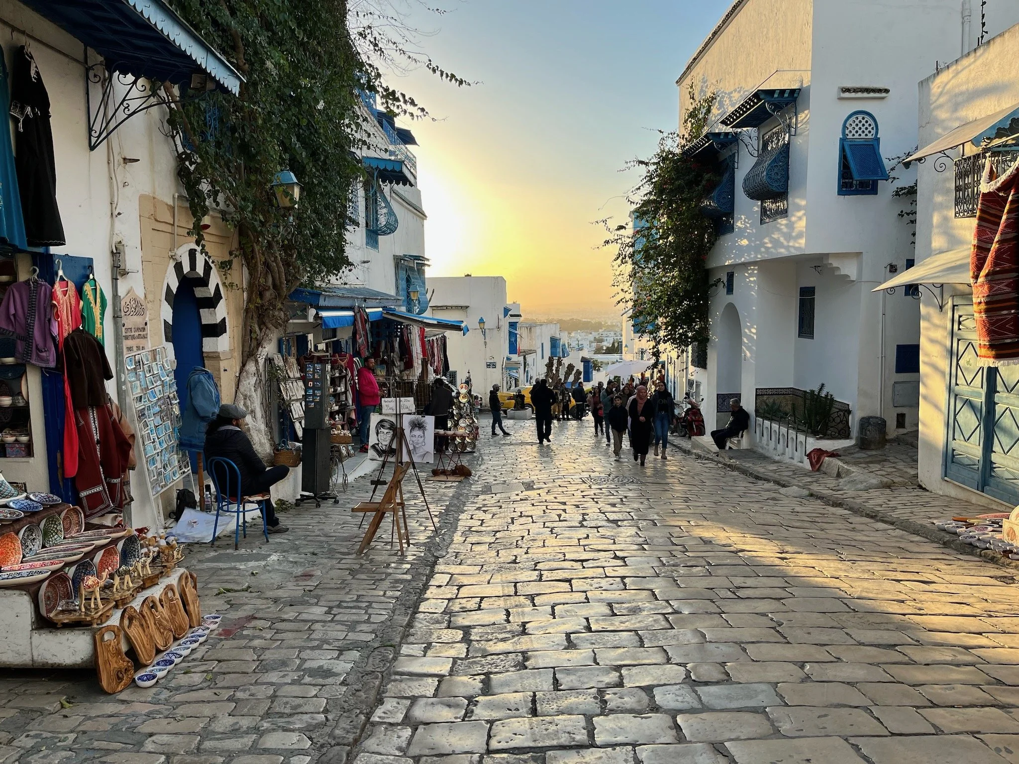 People walking and shopping on a cobblestone street lined with small shops selling souvenirs, with white buildings featuring blue accents, during sunset, in the Mediterranean city of Sidi Bou Said, Tunisia.