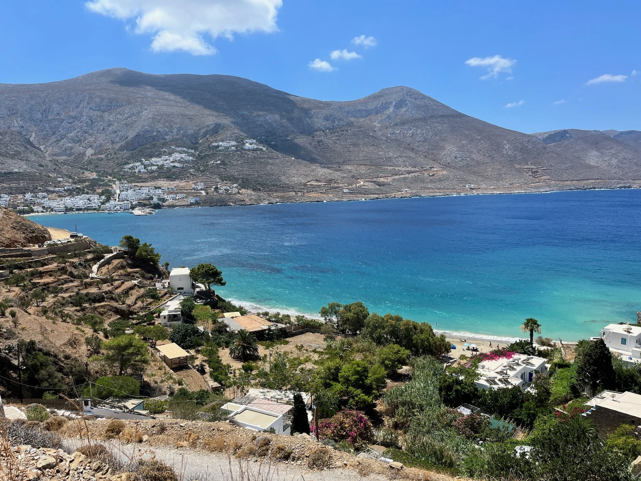 A coastal landscape of the Greek island Amorgos, in the Cyclades, with the turquoise water of the Aegean Sea, white buildings, and a mountain range in the background under a blue sky with some clouds.