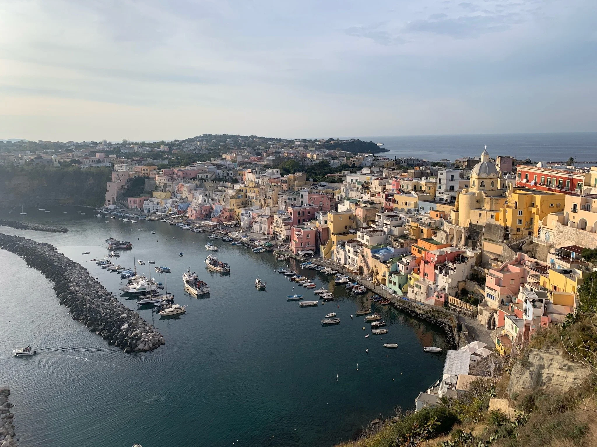 The picturesque town of Corricella on the island of Procida, in Campania, Italy,  with colorful buildings, a small harbor with boats, and a breakwater, situated on a hillside overlooking the ocean.