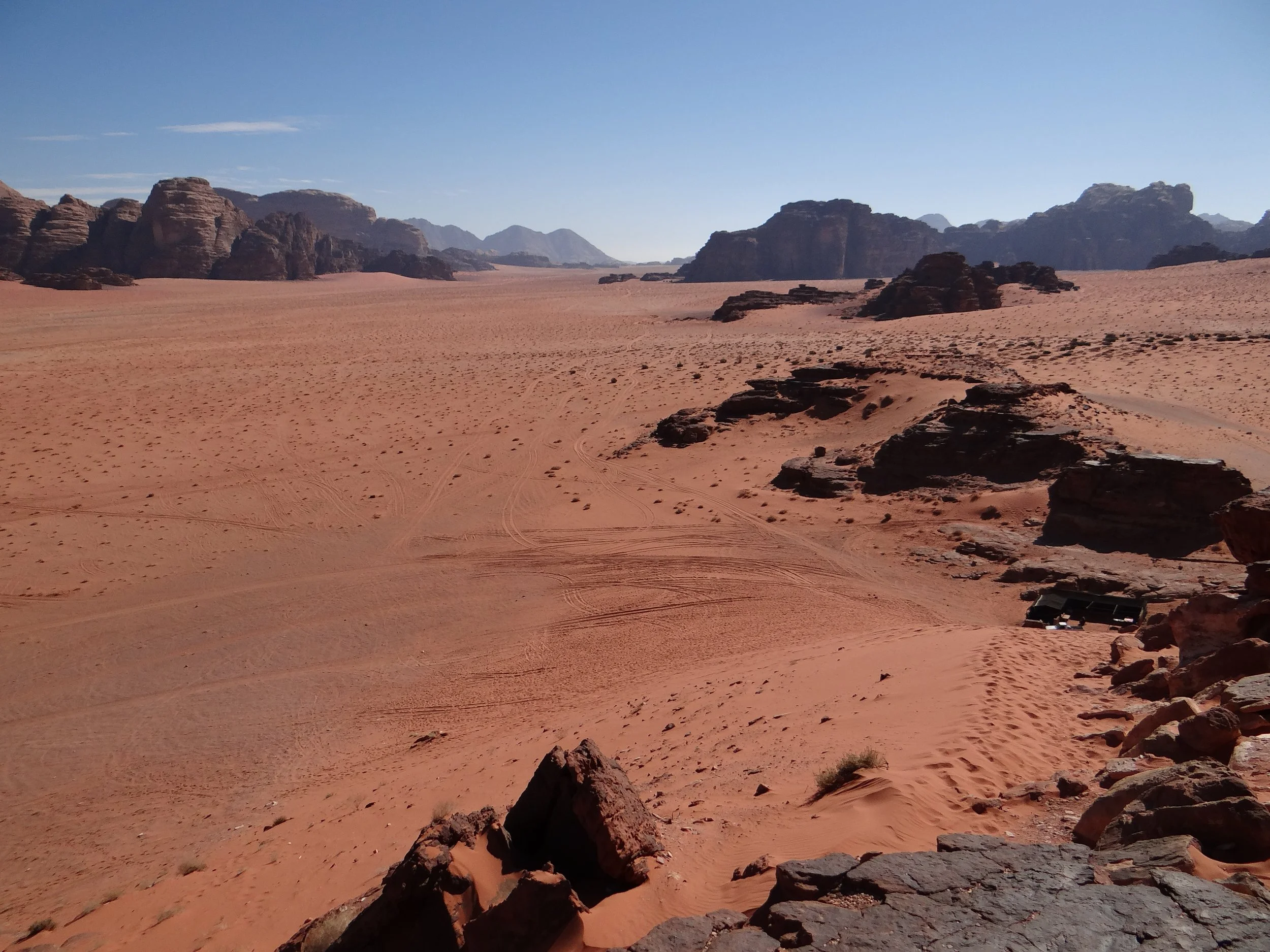 A vast desert landscape in the Wadi Rum protected area, in Jordan, with reddish sand, scattered rocks, distant rocky hill formations, and a clear blue sky.