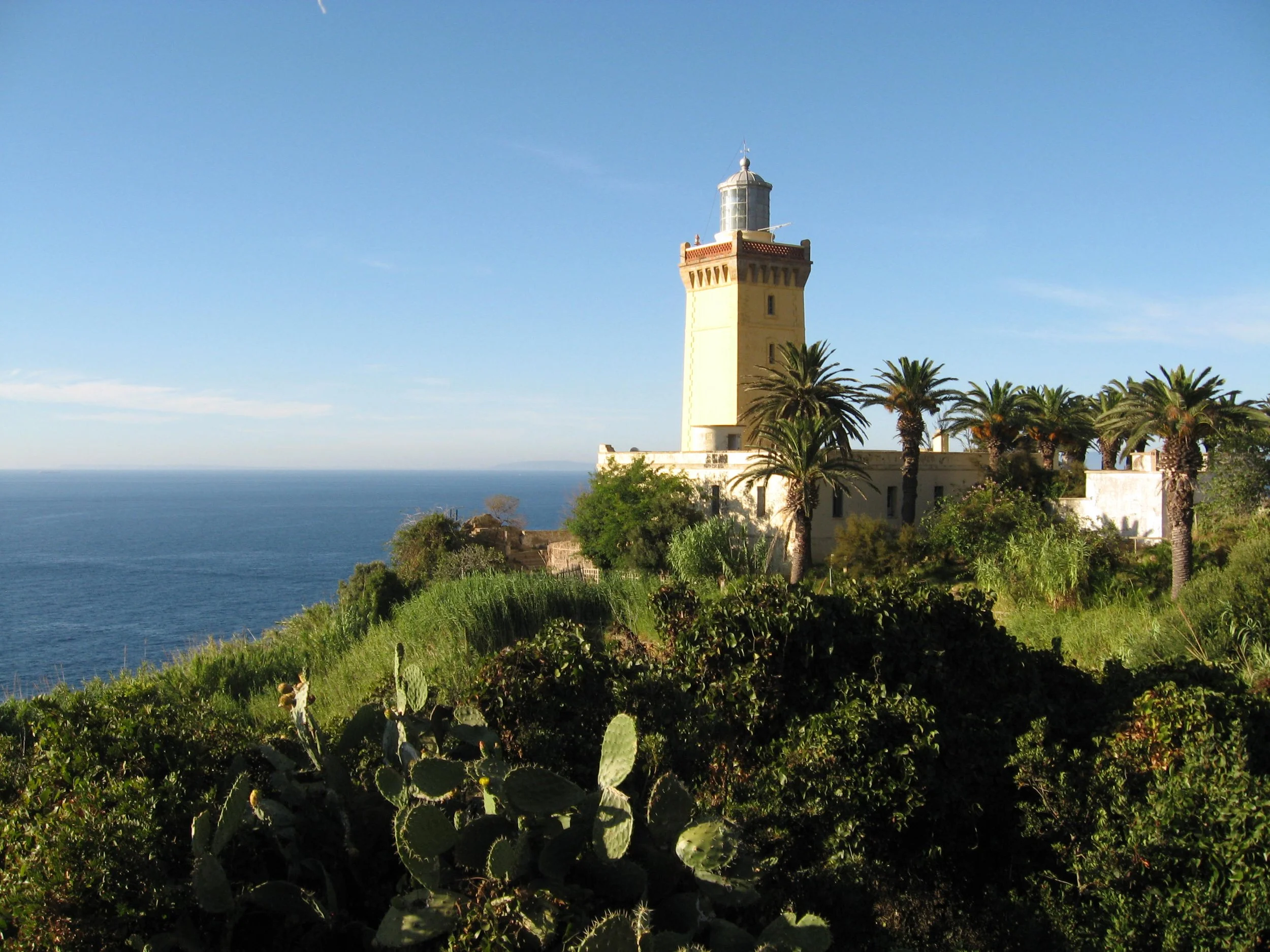 The lighthouse on a hill overlooking the ocean at Cap Spartel, Morocco, on the Strait of Gibraltar, surrounded by palm trees and lush green vegetation.